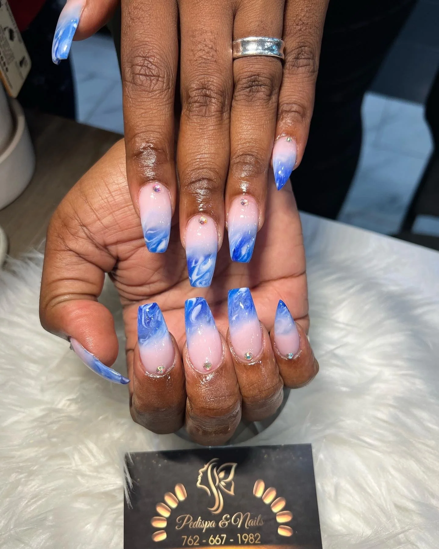 Close-up of a person's hands with long, blue and white gradient acrylic nails adorned with small rhinestones, and a silver ring on the middle finger.