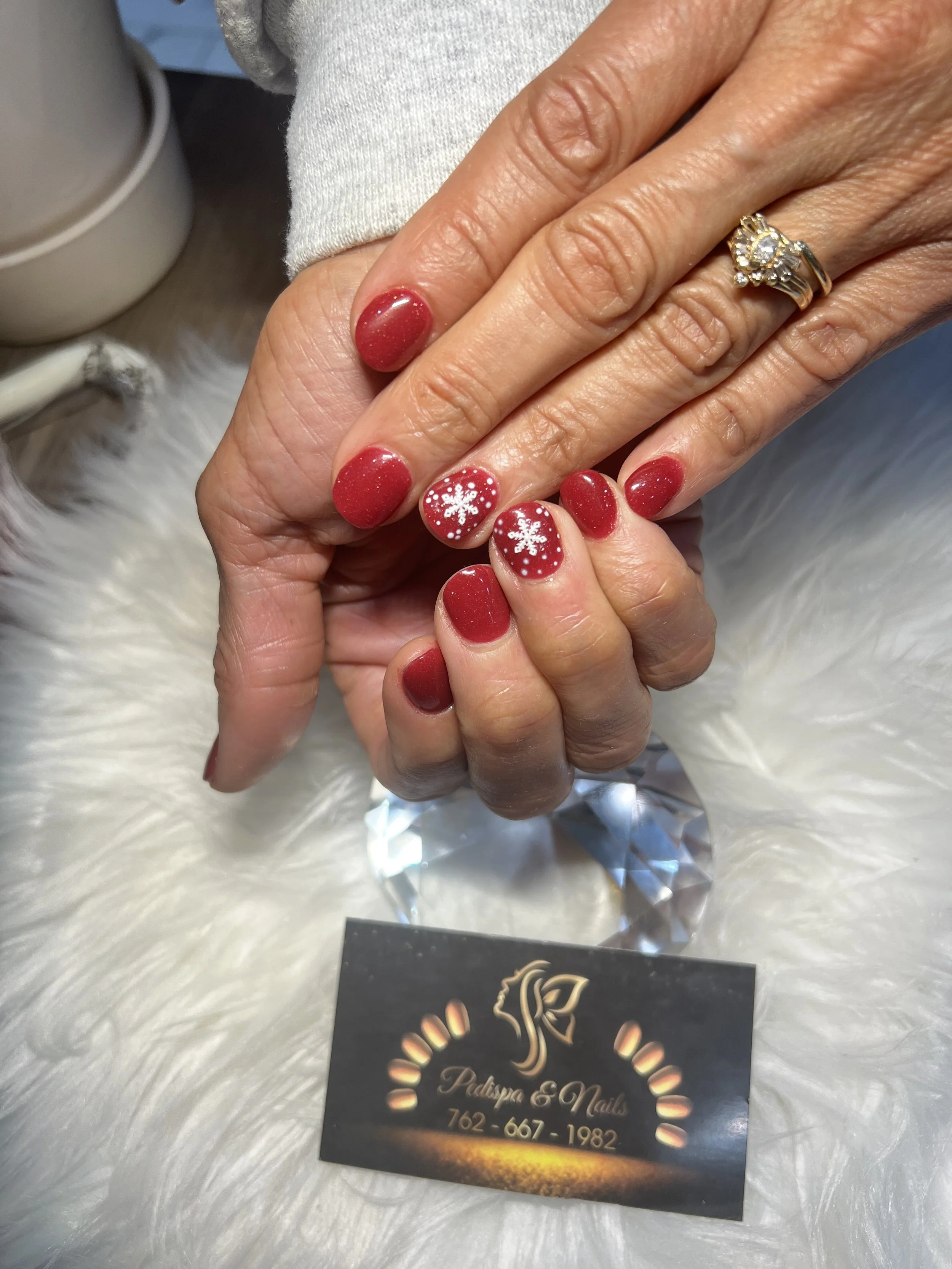 Close-up of a hand with red nail polish and white snowflake nail art, wearing a large ring, resting on a fluffy white surface with a business card for Palpape & Nails underneath. The background shows part of a container and a business card with conta