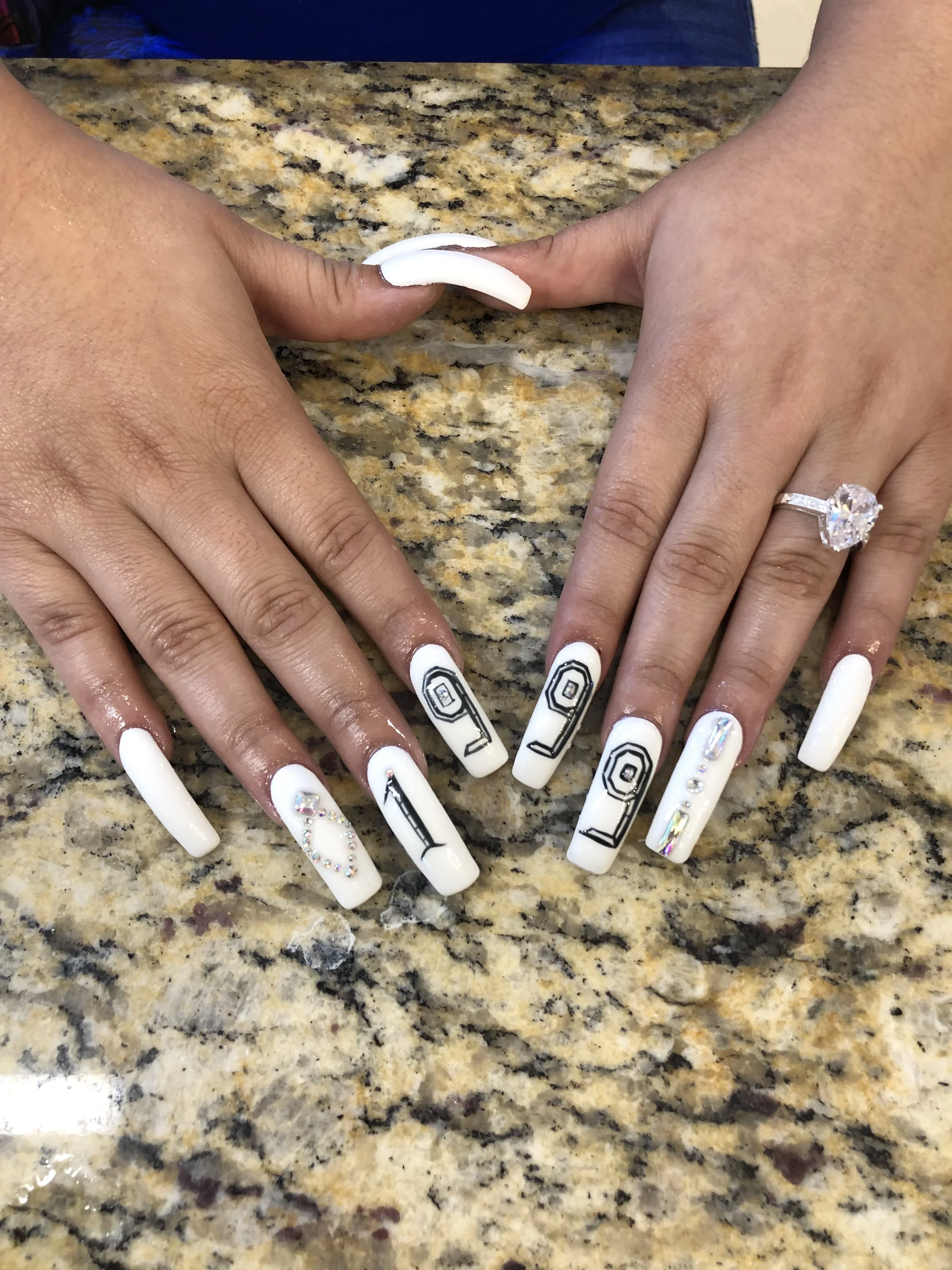 Hands with freshly painted white nails, some with black geometric designs, and an engagement ring with a large diamond, resting on a granite countertop.