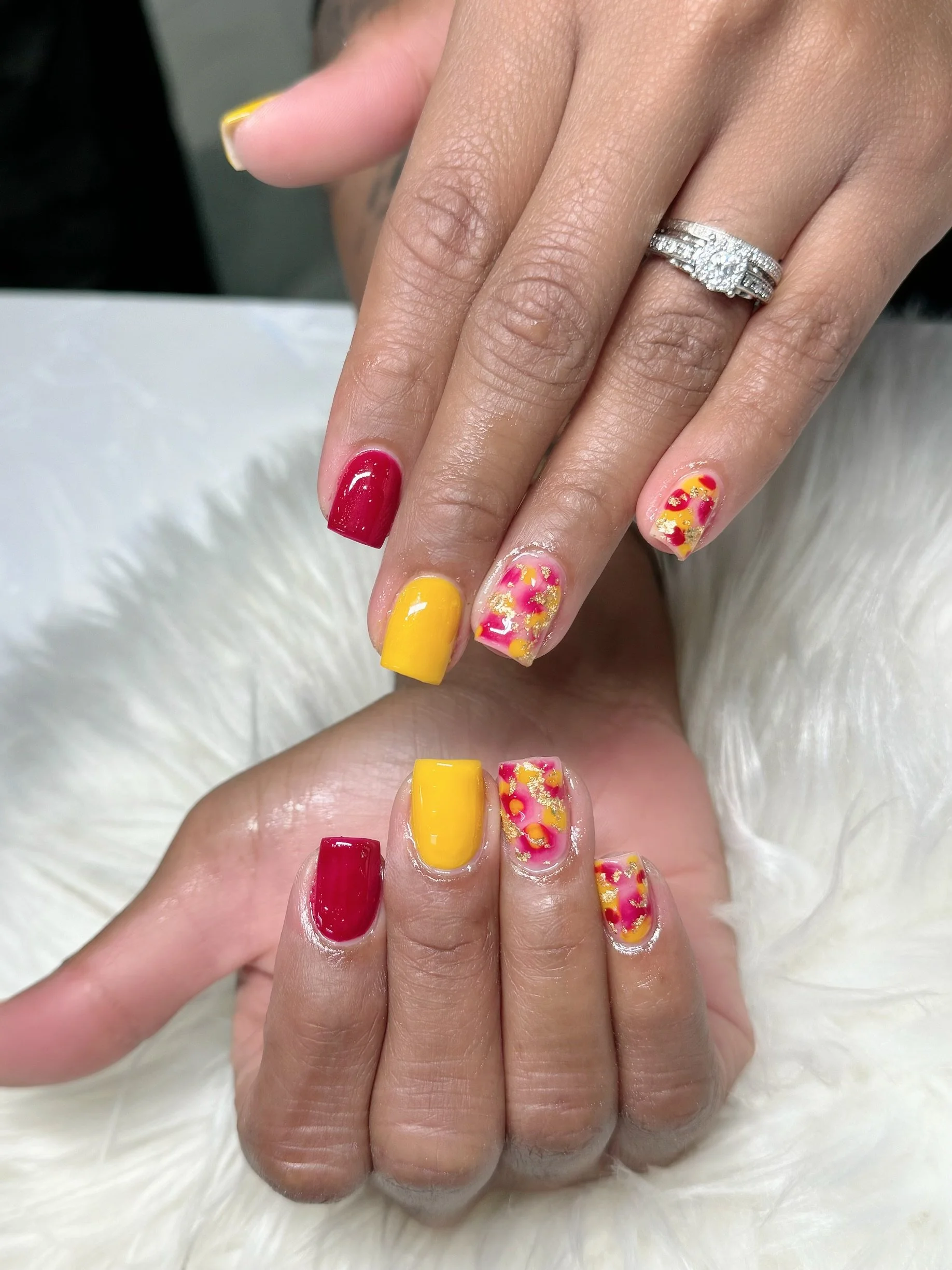 Close-up of a person's hand with colorful nail art, including yellow, red, and floral designs, wearing a diamond ring on the ring finger, against a white fluffy background.