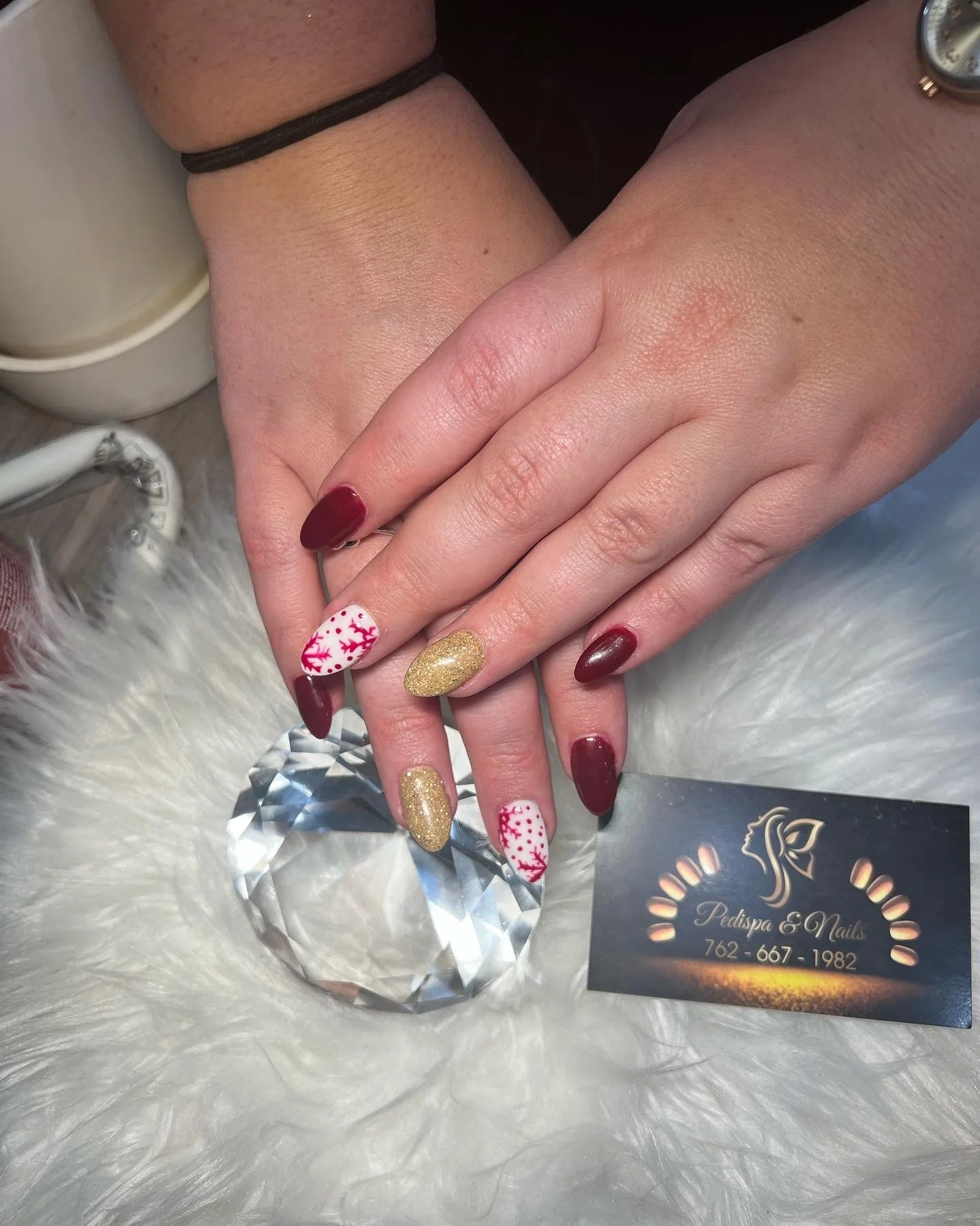 Manicure nails with seasonal red and white designs, gold glitter, and deep red polish, resting on a white furry surface with a business card for Pedispa & Nails.