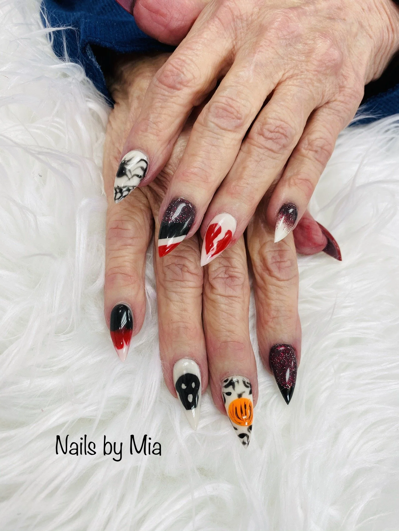 Close-up of a hand with Halloween-themed nail art on long, pointed nails featuring black and white marbled, red and white flames, black and white designs, and a pumpkin face, resting on a white furry surface with text that reads "Nails by Mia."