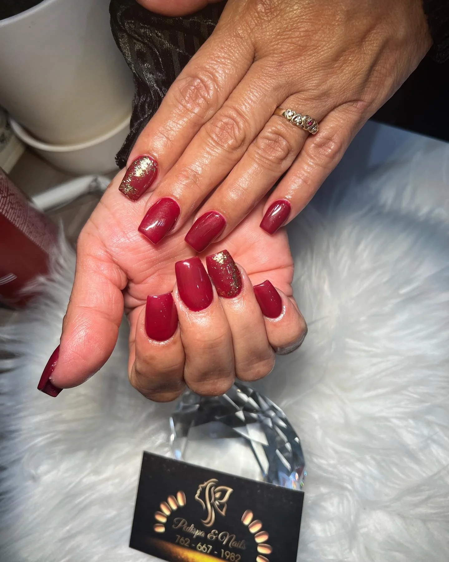 Close-up of a woman's hands with well-manicured red nails with gold glitter accents, wearing a silver ring with multiple pink and clear stones. The background includes a business card with a logo and contact information, and a blurred white furry sur