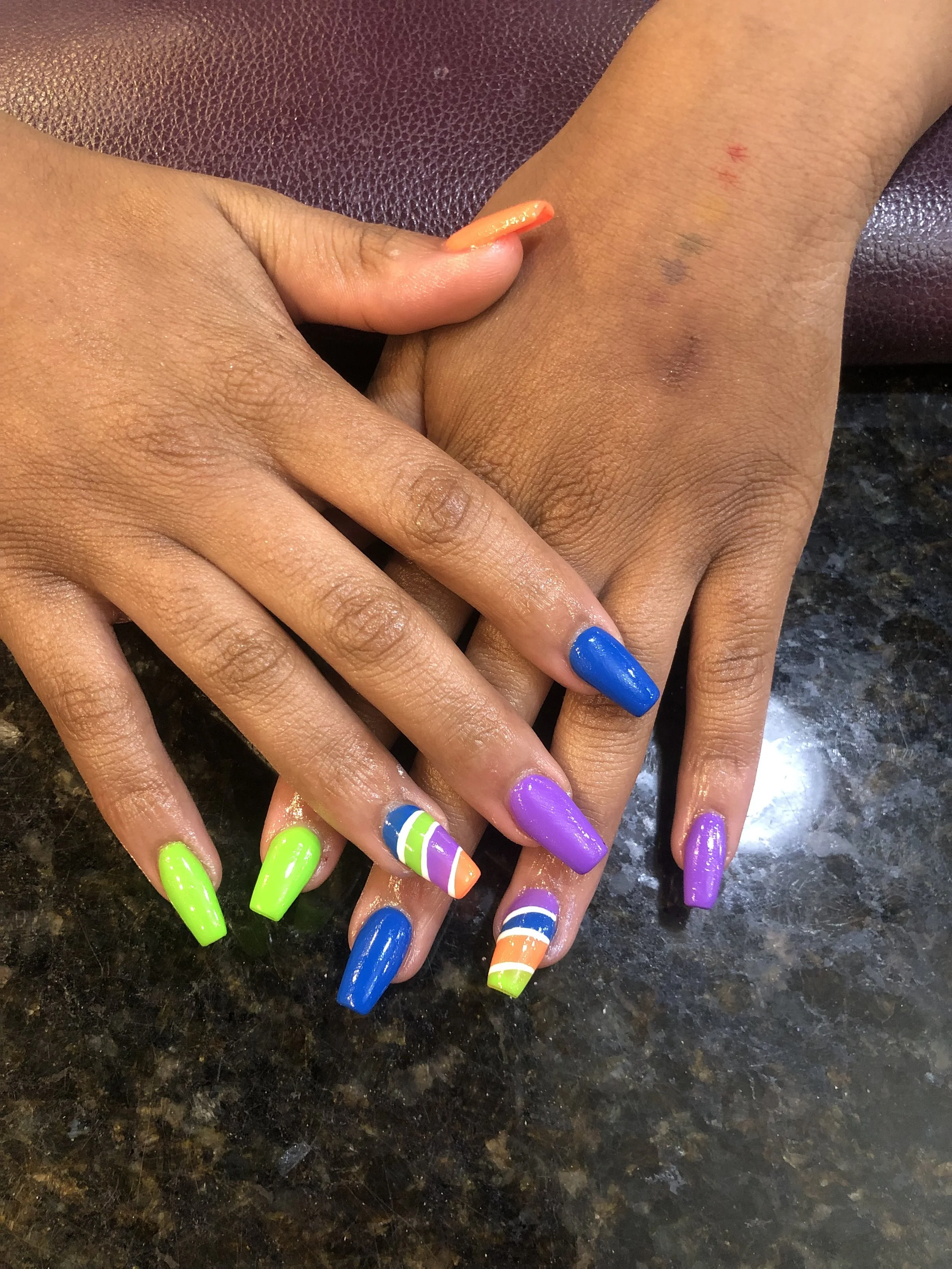 Multiple hands stacked together, each with brightly painted nails in neon green, blue, purple, and orange with white stripes, on a dark speckled surface.