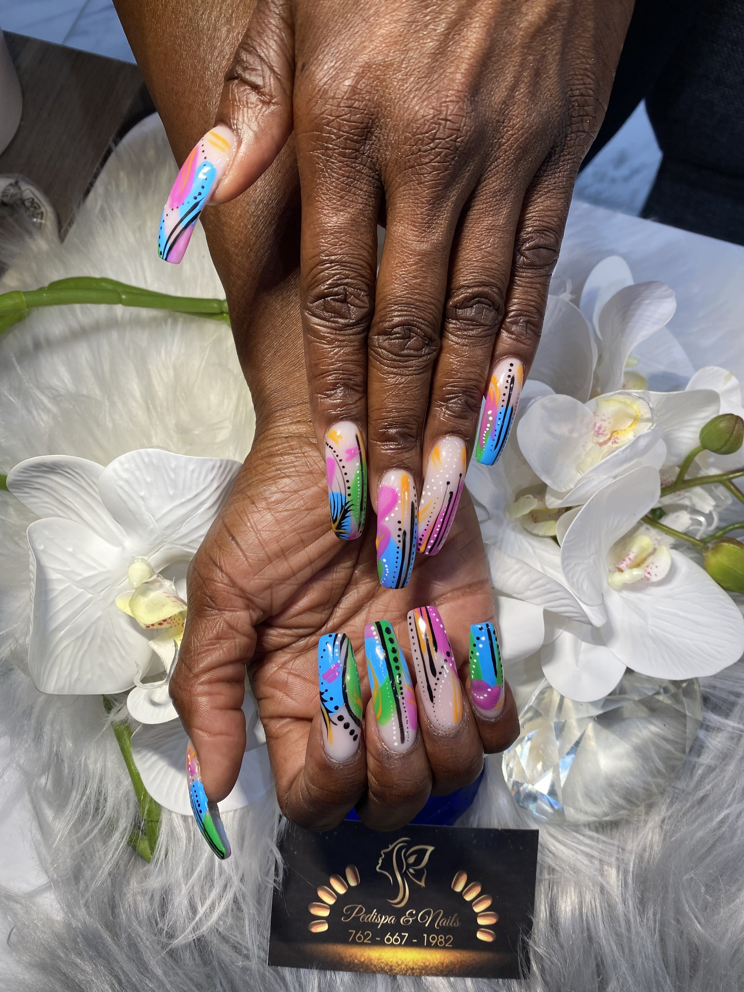 Close-up of hands with colorful, artistic nail designs, resting on a white orchid flower, with a business card for Pedispa & Nails in the background.