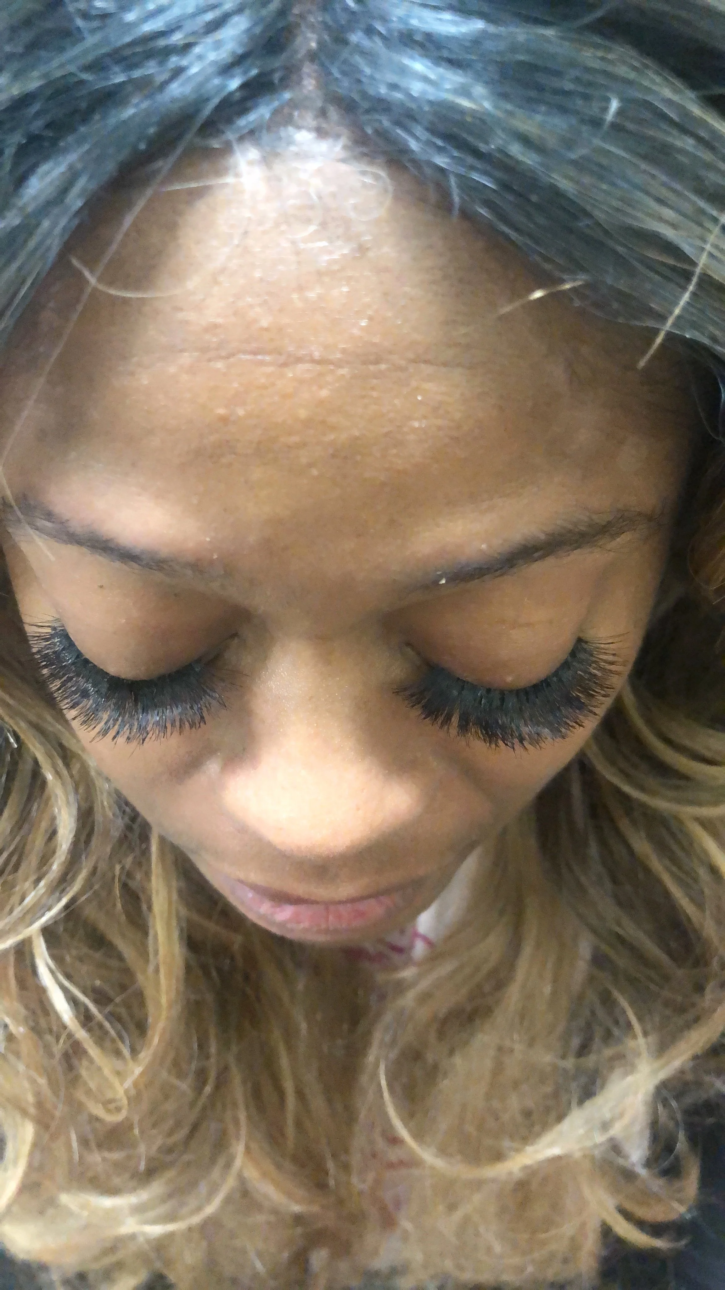Close-up of a woman's face with her eyes closed, showcasing long, dark eyelashes, and her forehead and hair partially visible with light brown and blonde curls.