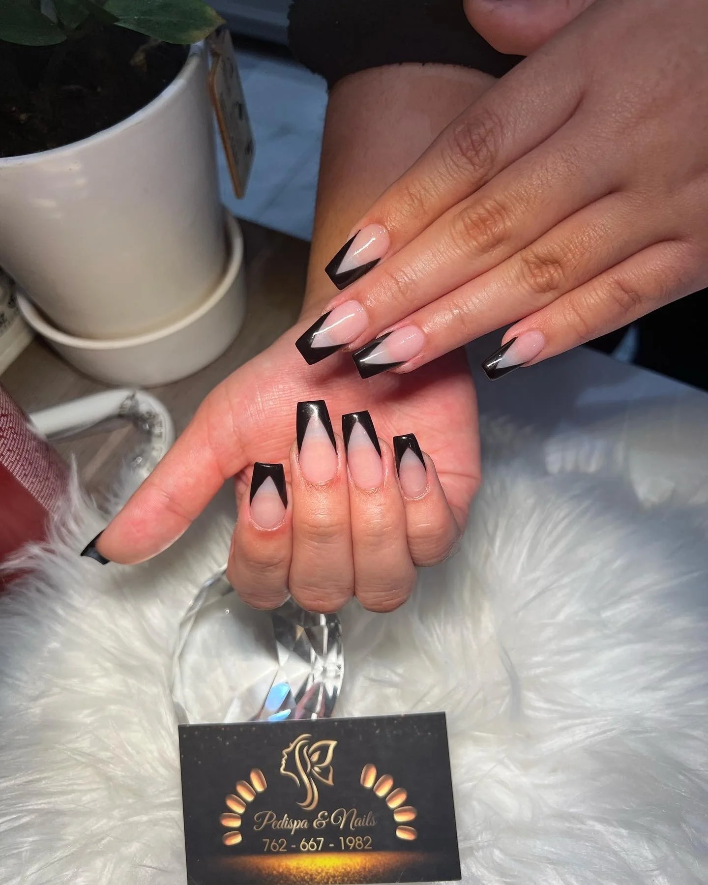 Close-up of a person's hands with freshly done black French tip acrylic nails, holding a small, shiny gemstone. In the background, there are stacked white ceramic cups and a potted plant. The person is sitting on a soft, white fur-like surface, and t