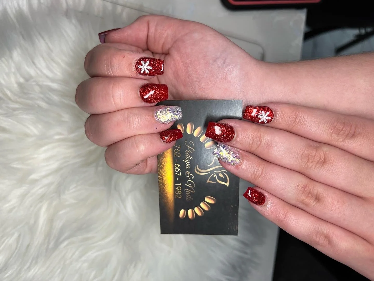 Close-up of hands with festive red, white, and iridescent glitter nail polish, holding a business card with gold lettering against a soft white furry background.