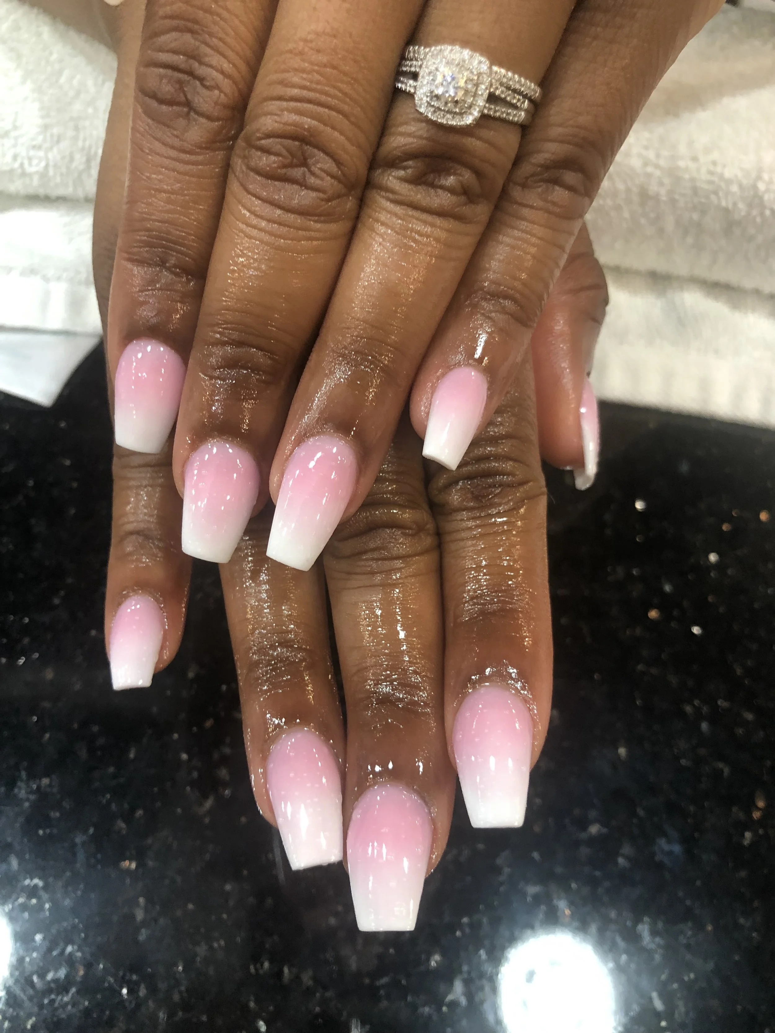 Close-up of a hand with a large diamond ring and acrylic nails that are light pink with a white tip, resting on a black granite countertop.