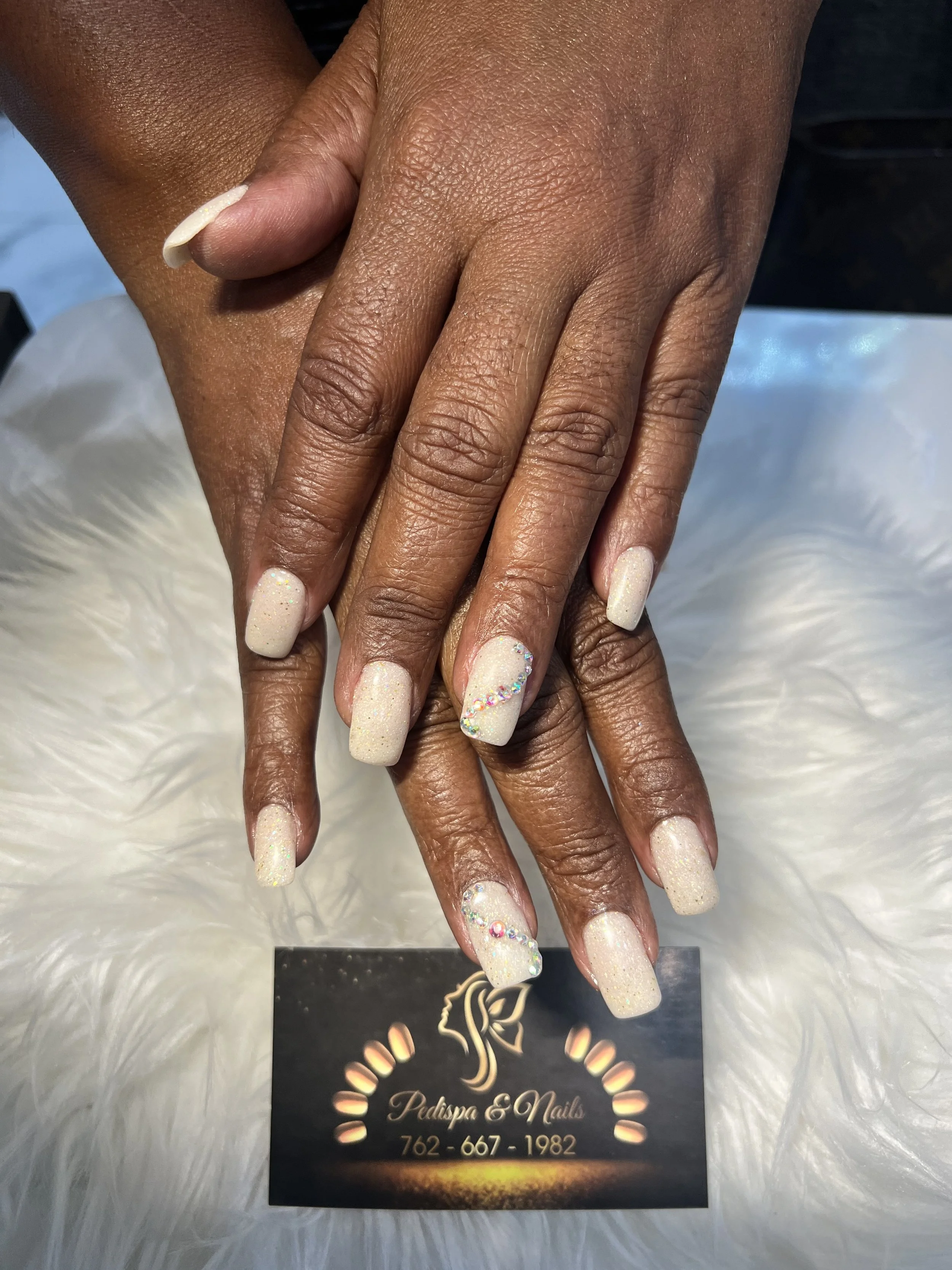 A pair of hands with well-manicured nails resting on each other. The nails are painted with a light, sparkly polish and decorated with colorful rhinestones at the tips. The hands are positioned on top of a black card with gold and white text and a lo