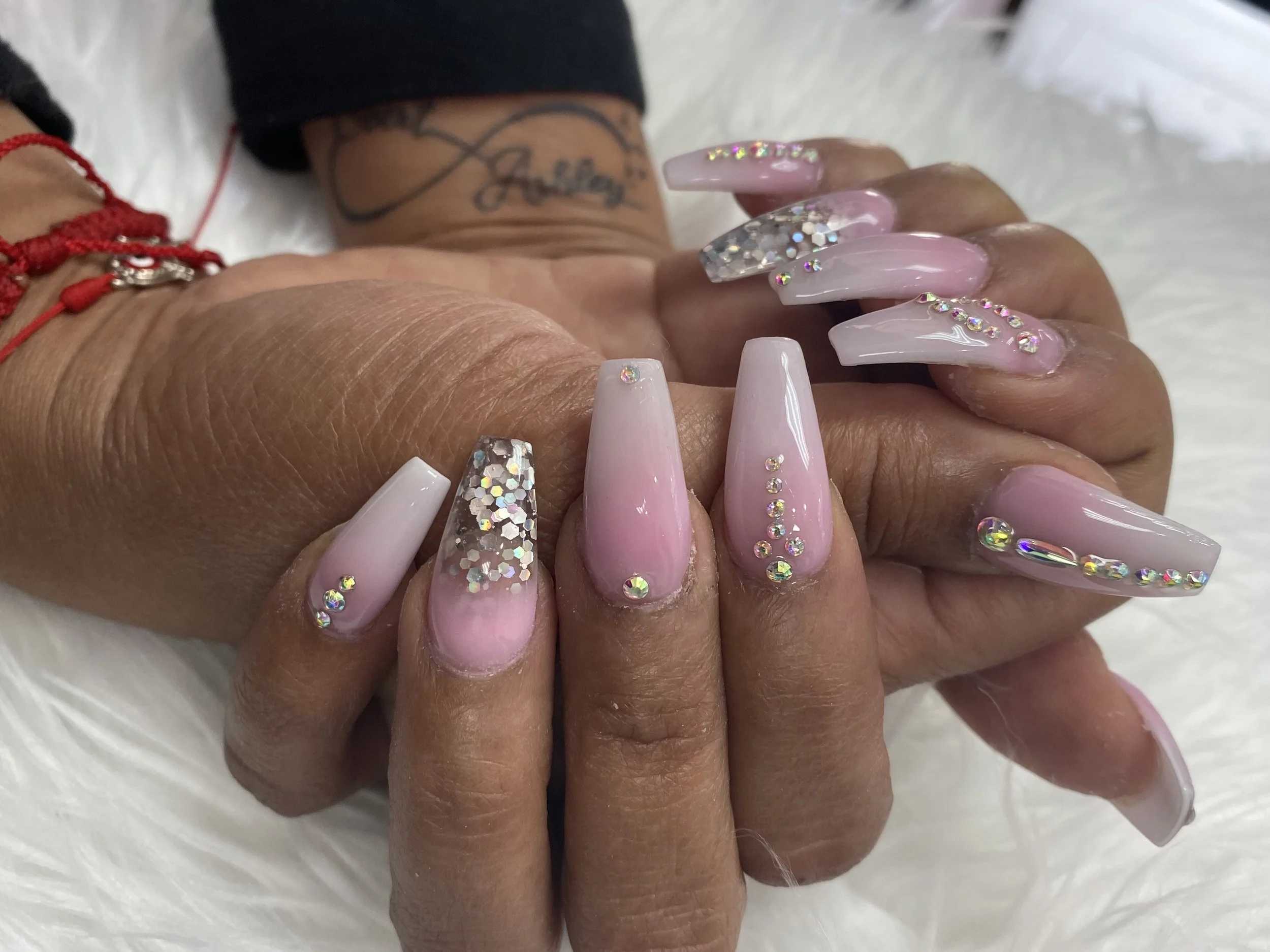 Close-up of a hand with long, pink acrylic nails decorated with rhinestones and glitter. The hand has a tattoo on the wrist and is wearing a red string bracelet.