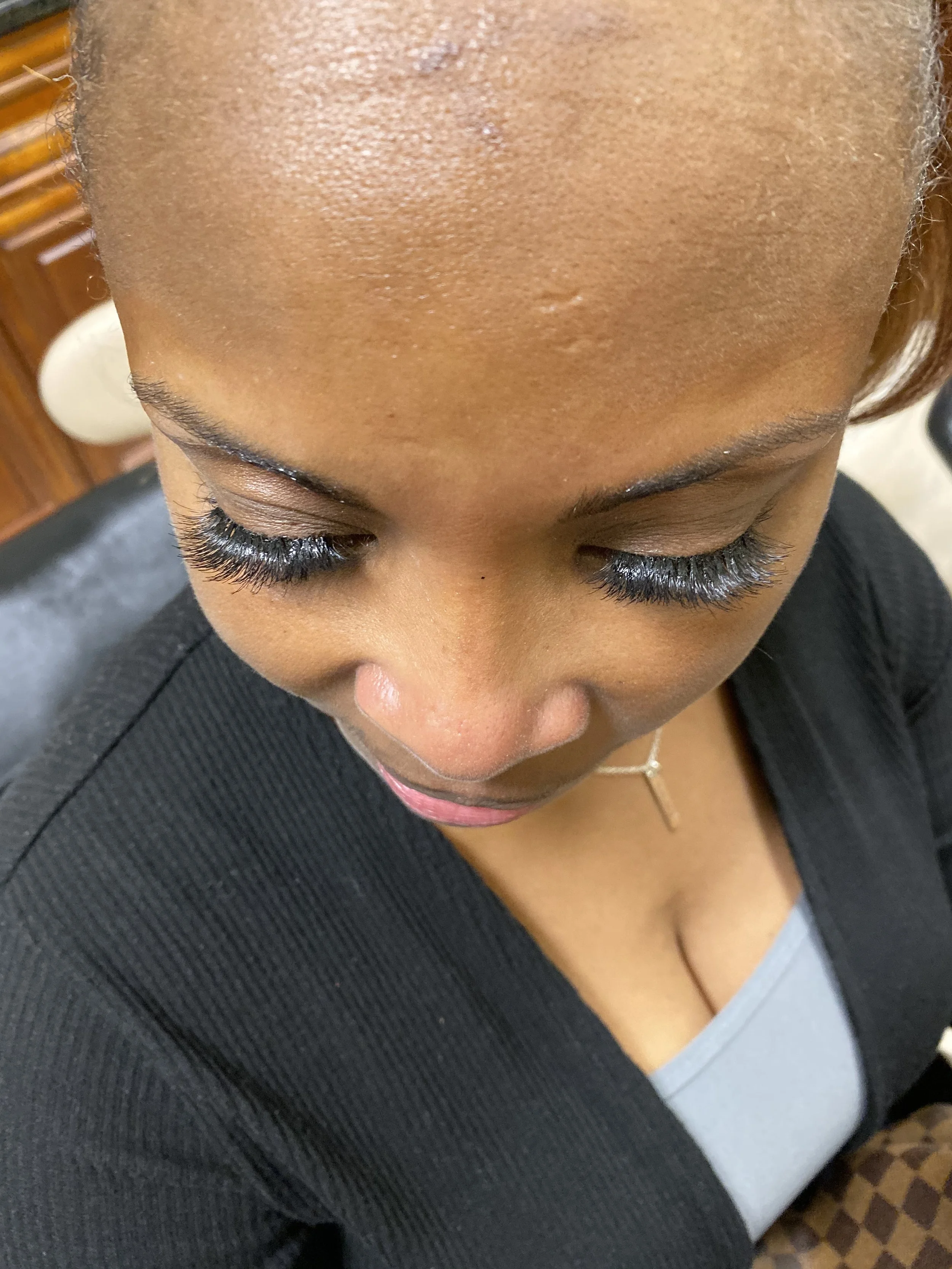 A person with short hair, closed eyes, and long eyelashes, wearing a black blazer over a gray top, with a gold necklace, sitting in a chair in what appears to be a salon or similar setting.