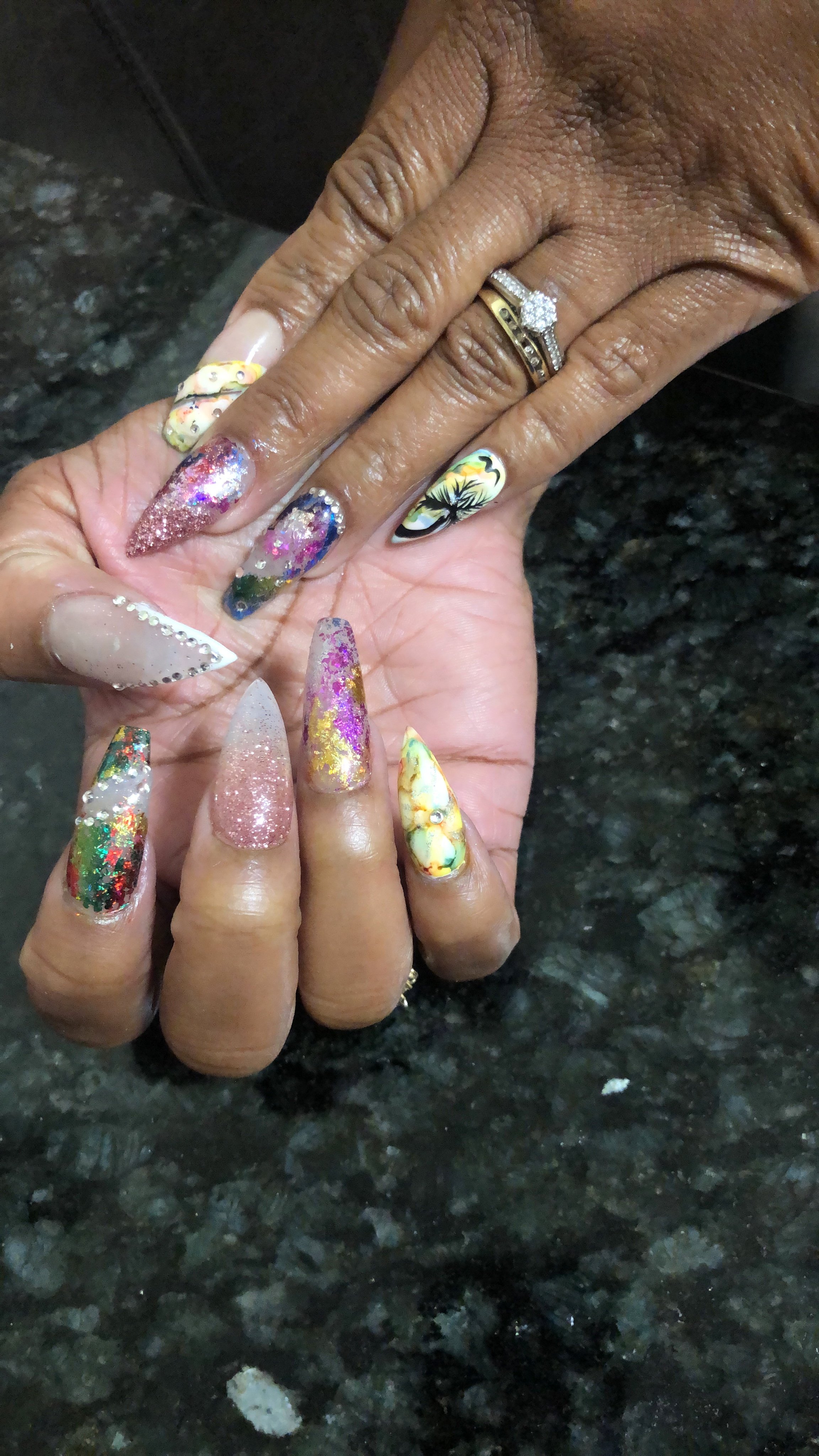 Close-up of two hands with decorated nails and rings. The nails are colorful with various designs, and the rings are gold and silver, one with a large gemstone.