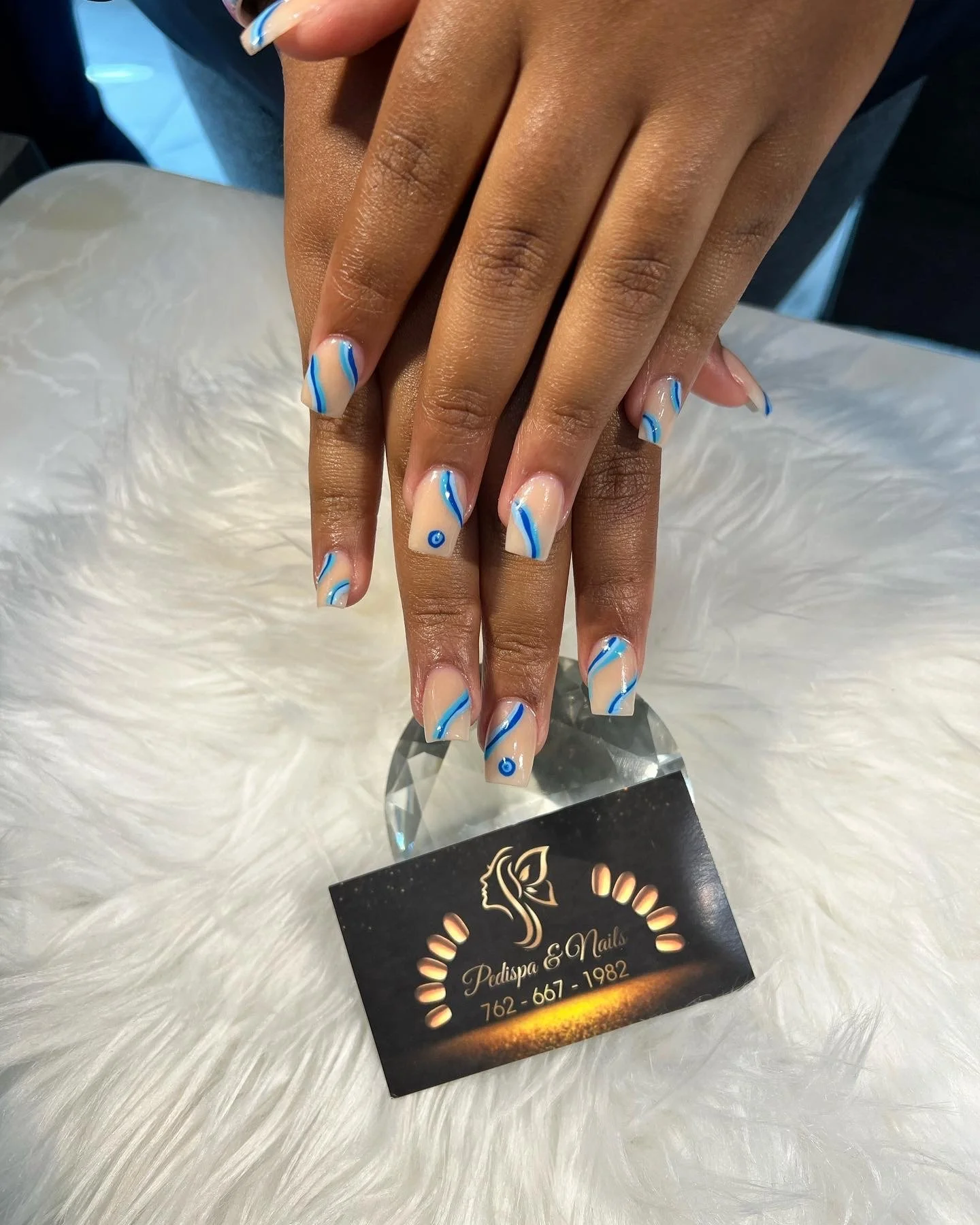 Close-up of a woman's hands with freshly done blue and white swirl acrylic nails, resting on a faux fur surface. There is a business card for Pedispa & Nails visible in the image.