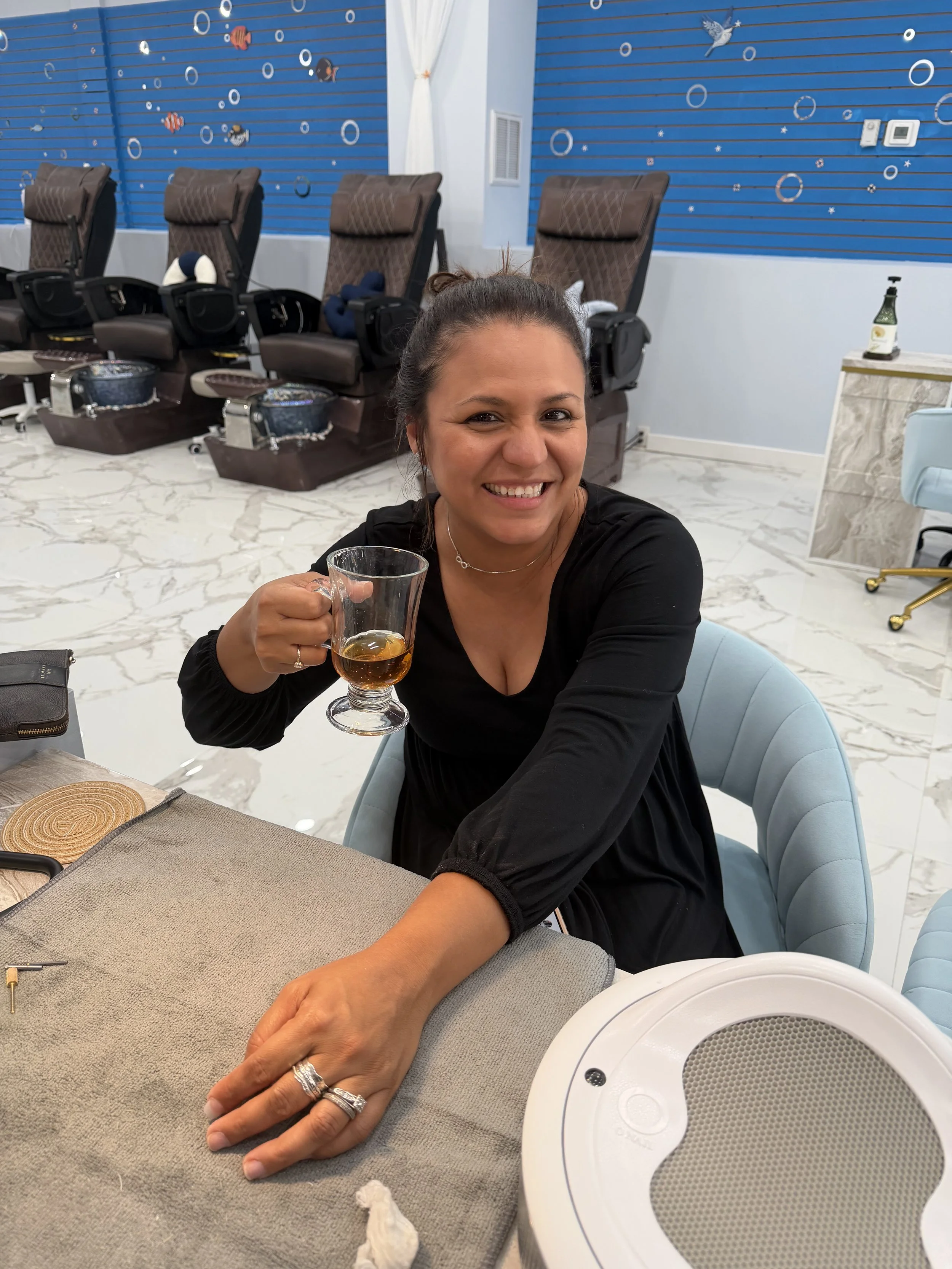 Woman smiling, holding a glass of whiskey, seated at a table with a beige placemat and silverware, in a room with decorated blue walls and chairs, with a white speaker in the foreground.
