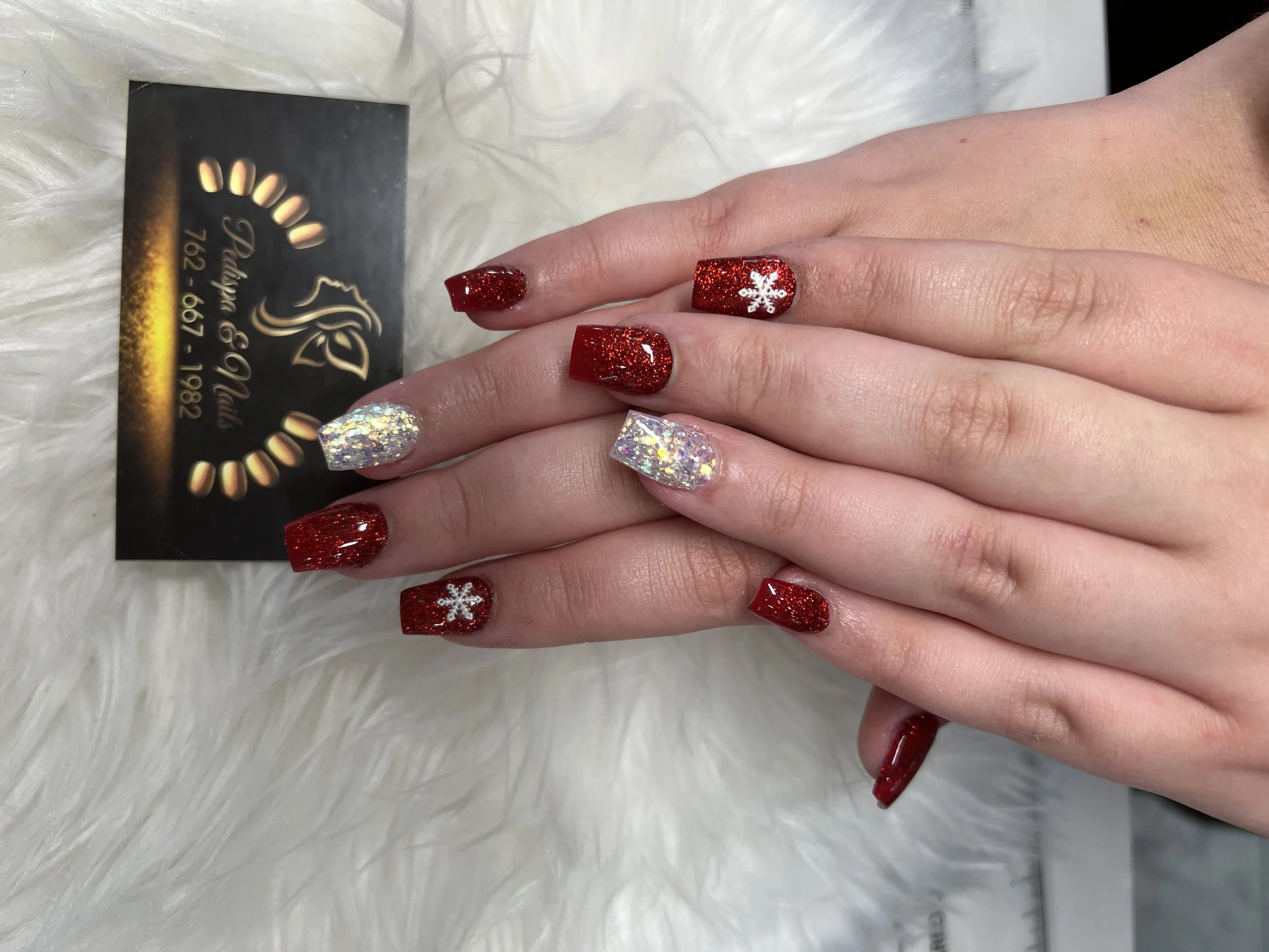 Close-up of a hand with red glitter nail polish and a white snowflake design on one nail, resting on a fluffy white surface, with a business card for a nail salon nearby.