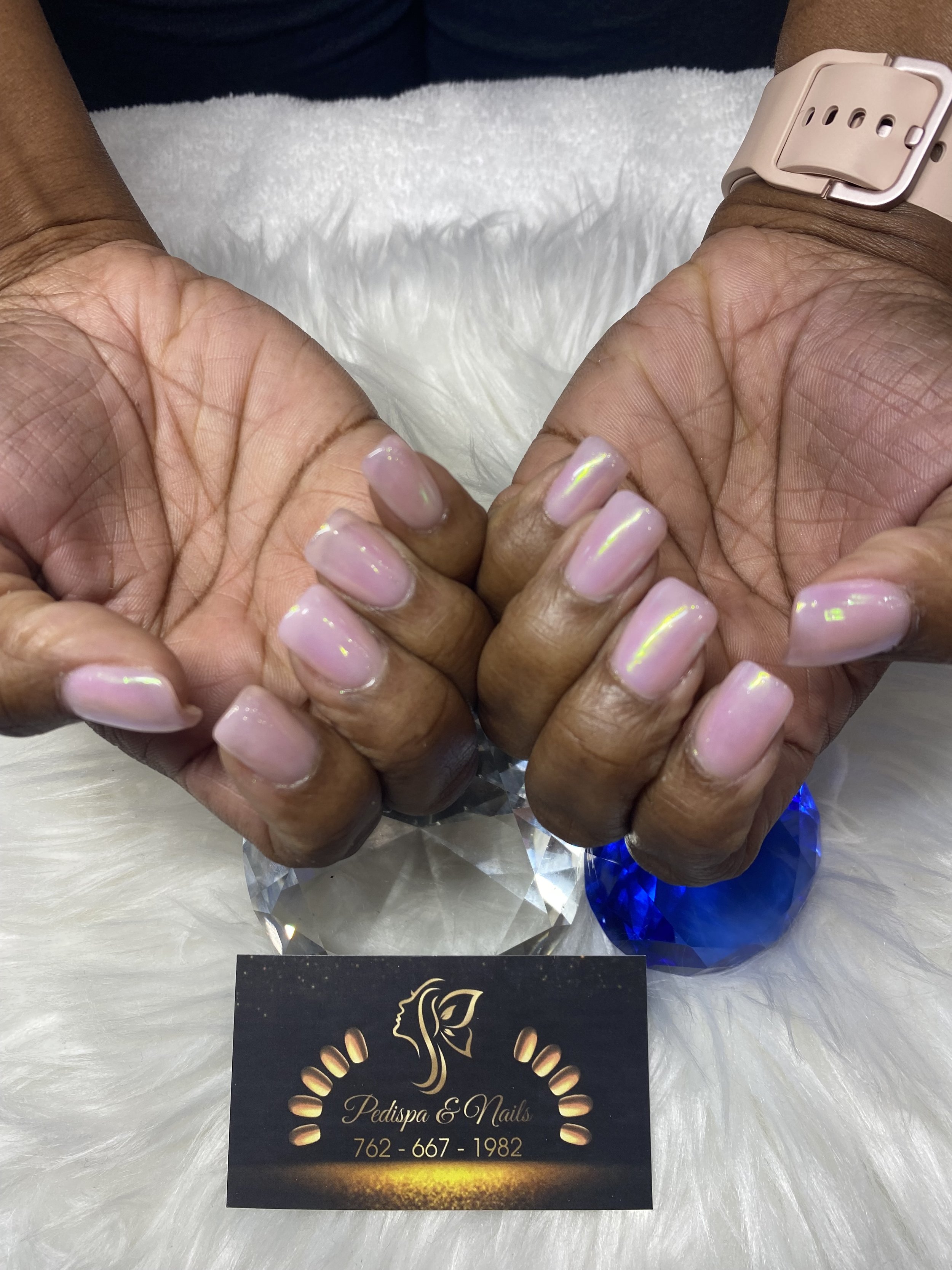 Close-up of hands with manicured nails holding a business card for Pedispa & Nails. The card features a woman's face silhouette surrounded by golden dots, and includes a phone number.