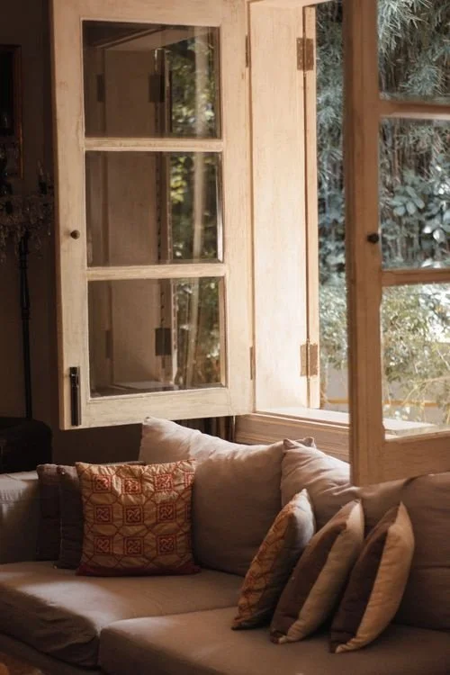 Open window with wooden frames and glass panes, revealing greenery outside, above a cozy beige couch with decorative pillows inside a room.