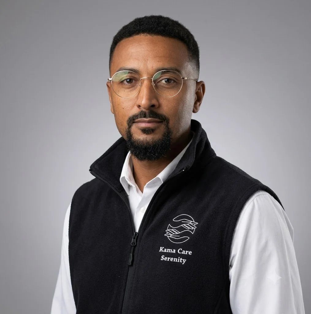 A man with glasses, a goatee, and short curly hair, wearing a black vest with a logo and text, over a white collared shirt, against a plain gray background.