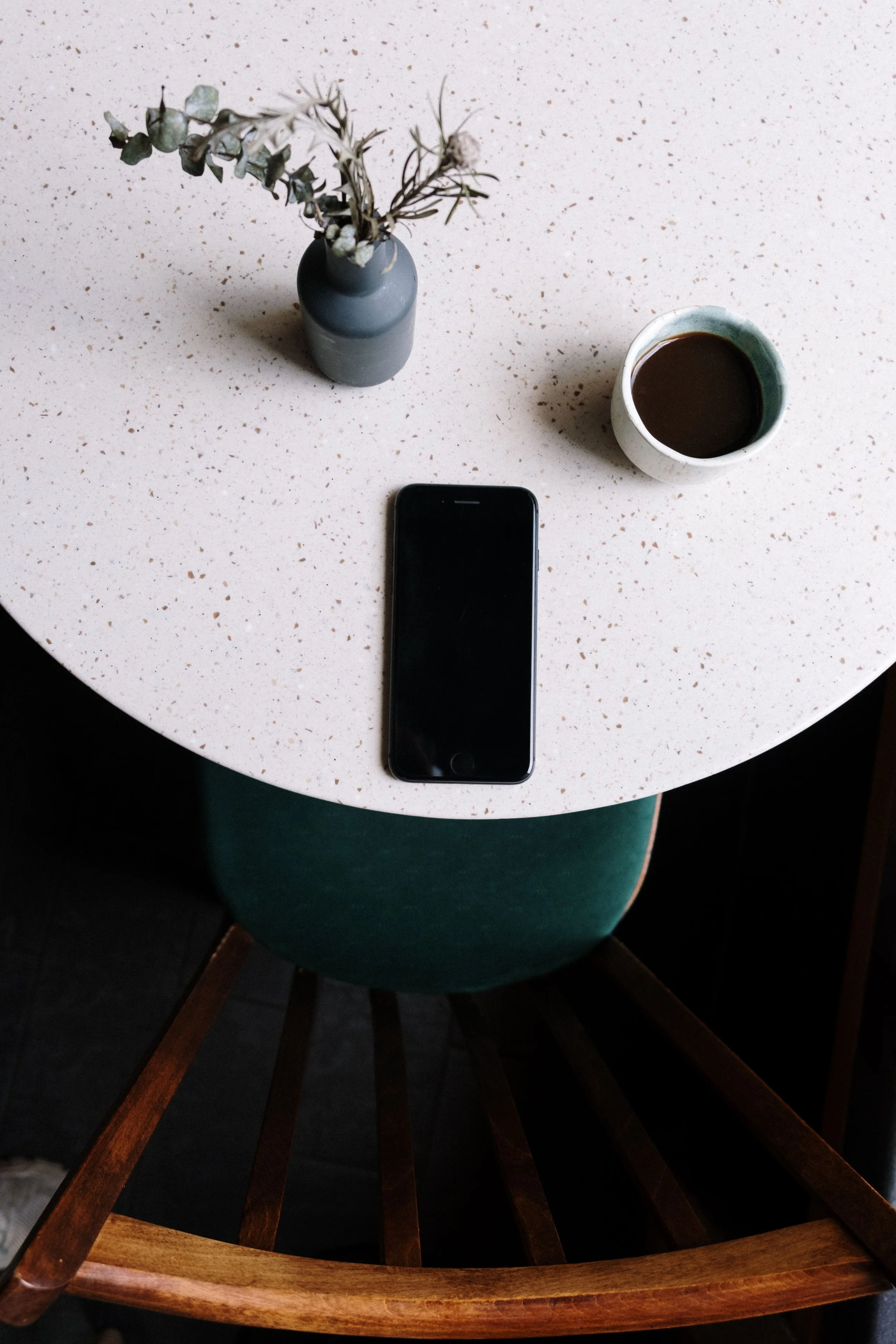 A round speckled beige table with a dark-colored vase with dried leaves and flowers, a cup of dark coffee, and a black smartphone on top. The table is above a wooden chair with green upholstery.
