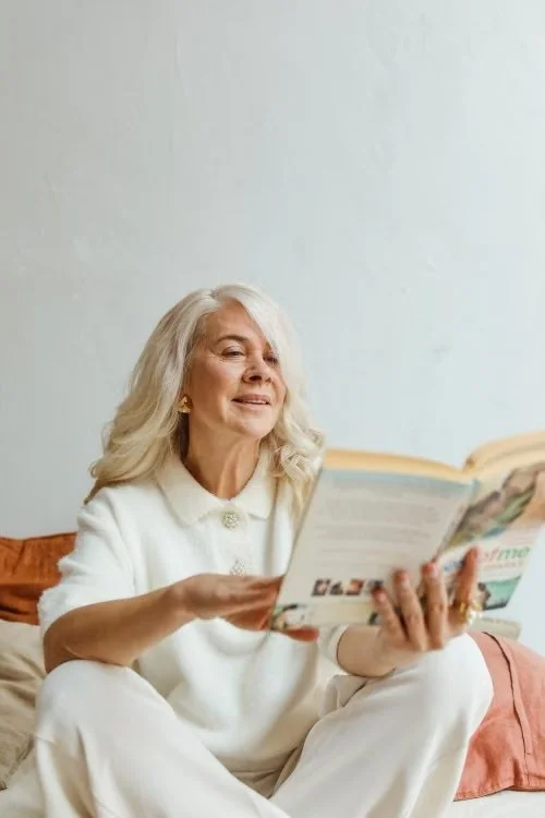 Older woman with long gray hair sitting on a couch, smiling and reading a magazine.