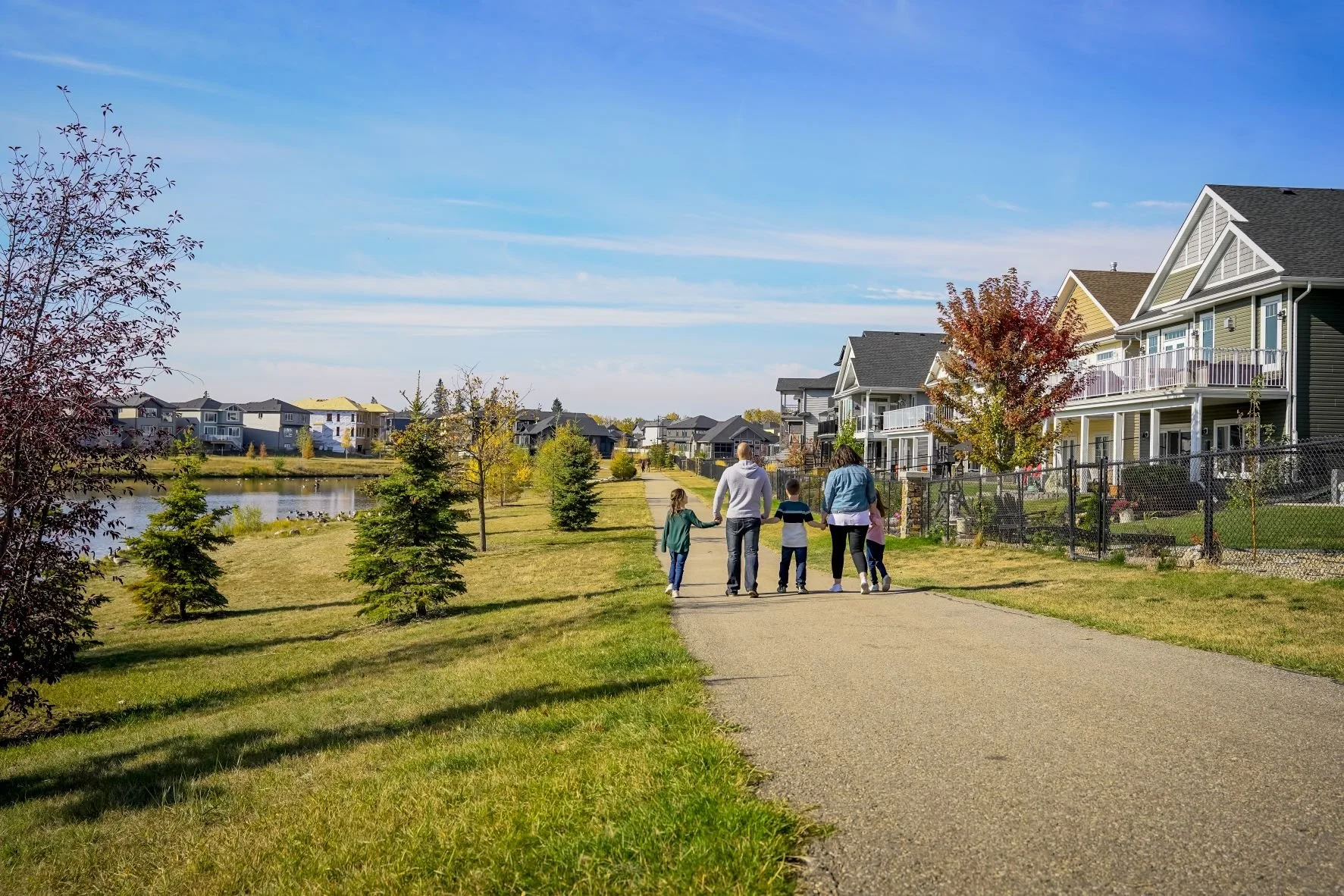 A family of five walking along a paved path next to a pond in Beaumont. The sky is blue with scattered clouds, and houses with porches. Trees with fall foliage line the path near Celeste at Azur townhome rentals in Beaumont.