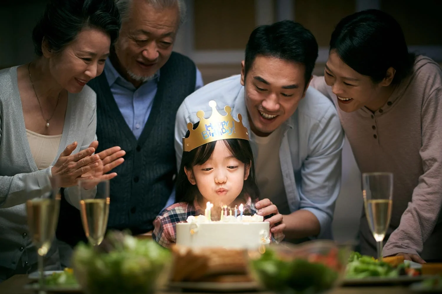Family gathered around a young girl with a birthday cake, wearing a ‘Happy Birthday’ crown, blowing out candles, with candles and food on the table.