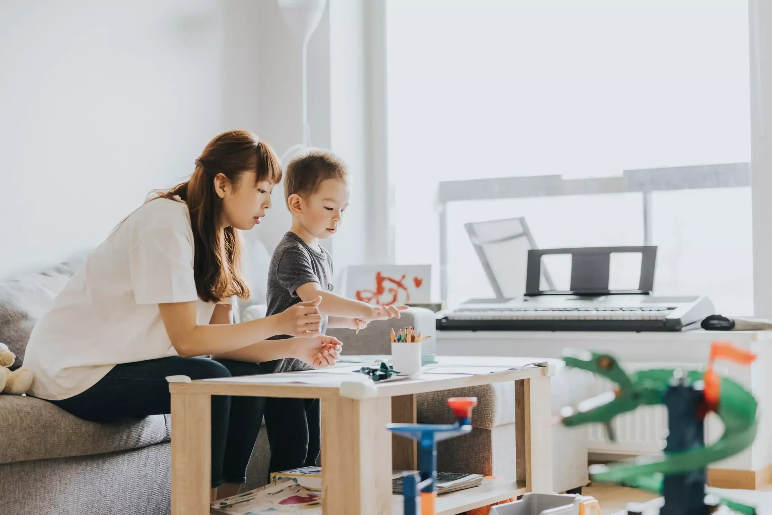 A woman and a young boy sitting at a table, engaging in arts and crafts. The woman is teachinghe boy with his activity. The background includes a keyboard and large windows letting in natural light at Celeste at Azur townhome rentals in Beaumont.
