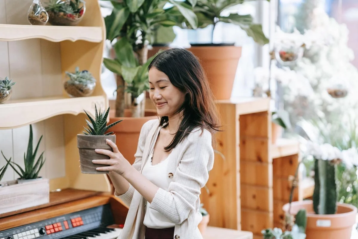 A woman with long dark hair holding a potted succulent plant in a greenhouse or plant shop, surrounded by various potted plants and greenery.