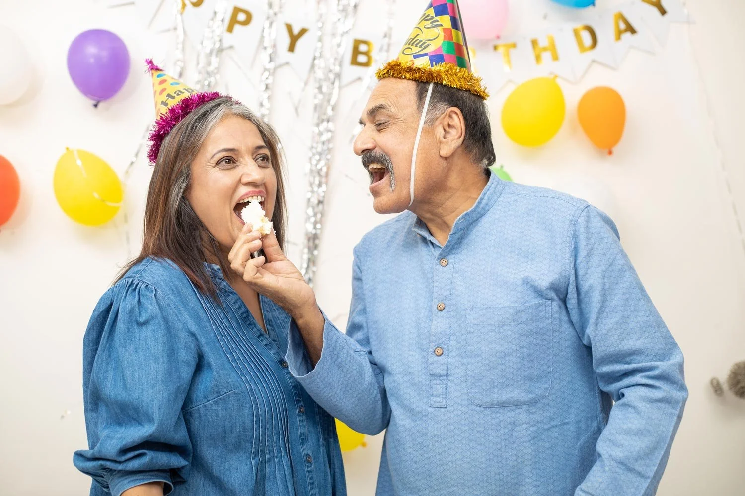 A man and woman at a birthday party wearing colorful party hats, surrounded by balloons and decorations. The man is feeding the woman cake, and they are both smiling and laughing.