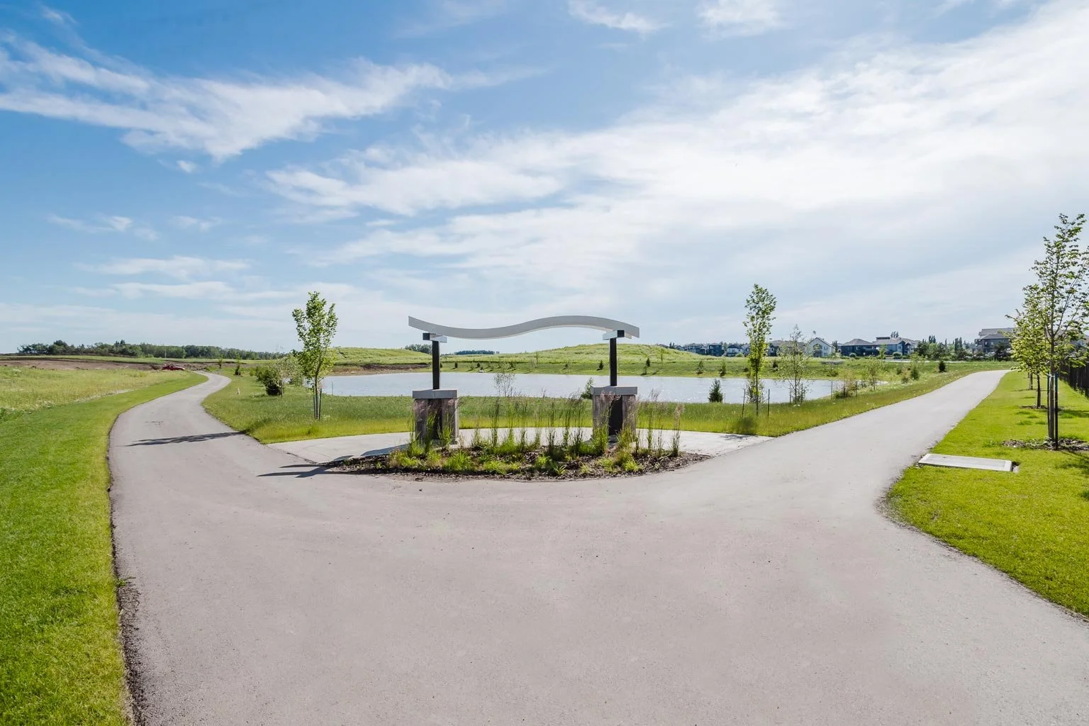 A park with a split pathway, a water body, and a decorative structure in the center, with trees and residential buildings in the background under a partly cloudy sky, located near Celeste at Azur townhome rentals in Beaumont.