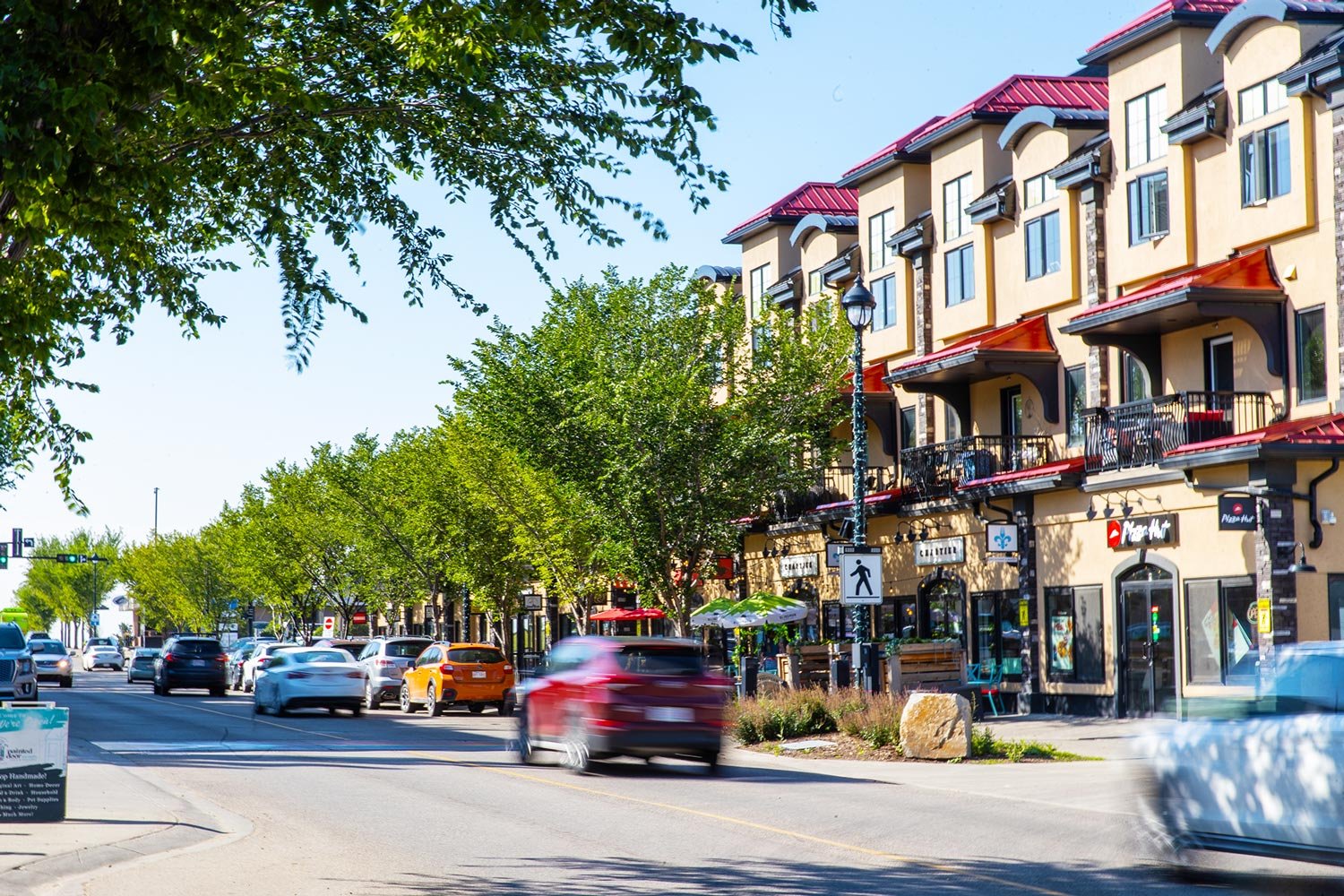 A busy street with multiple cars driving past a row of trees and multi-story commercial buildings with red roofs and balconies, sidewalk cafes, and signboards, located in Beaumont and featured for Celeste at Azur townhome rentals.