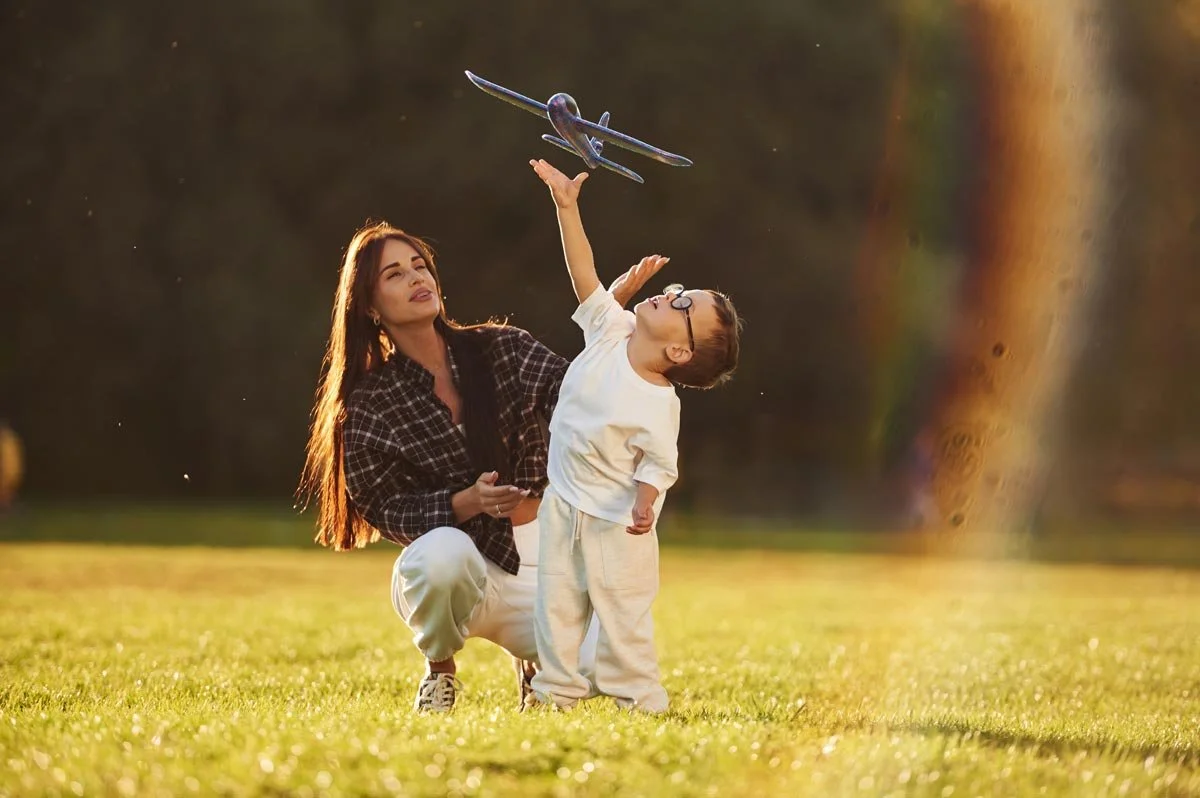 A woman and a young boy flying a toy airplane outdoors during sunset.