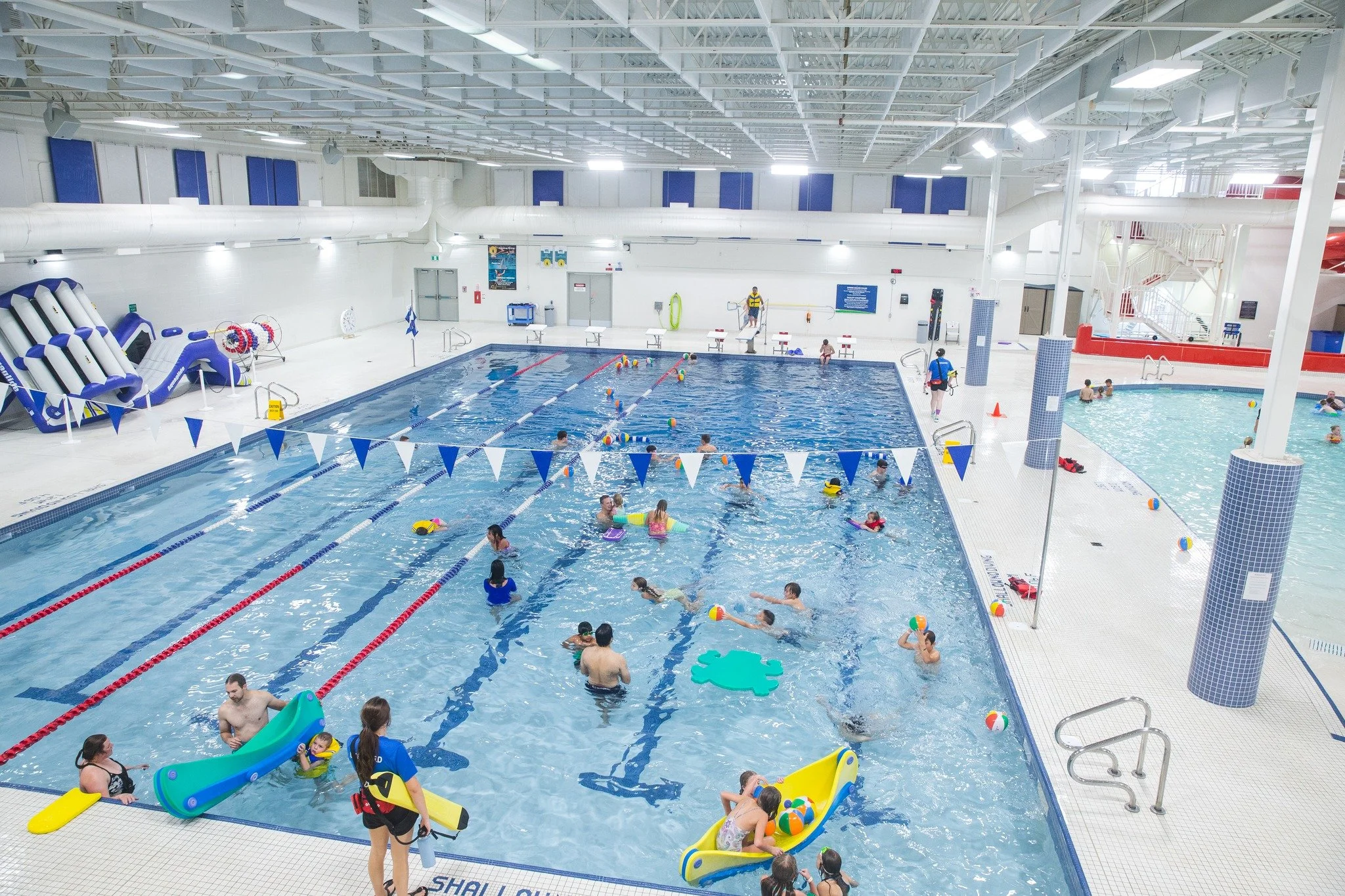 Indoor community swimming pool with children and adults swimming and playing. Pool has lanes, colorful floaties, and beach balls. Lifeguards oversee from outside the pool. The ceiling has white beams and bright lighting.