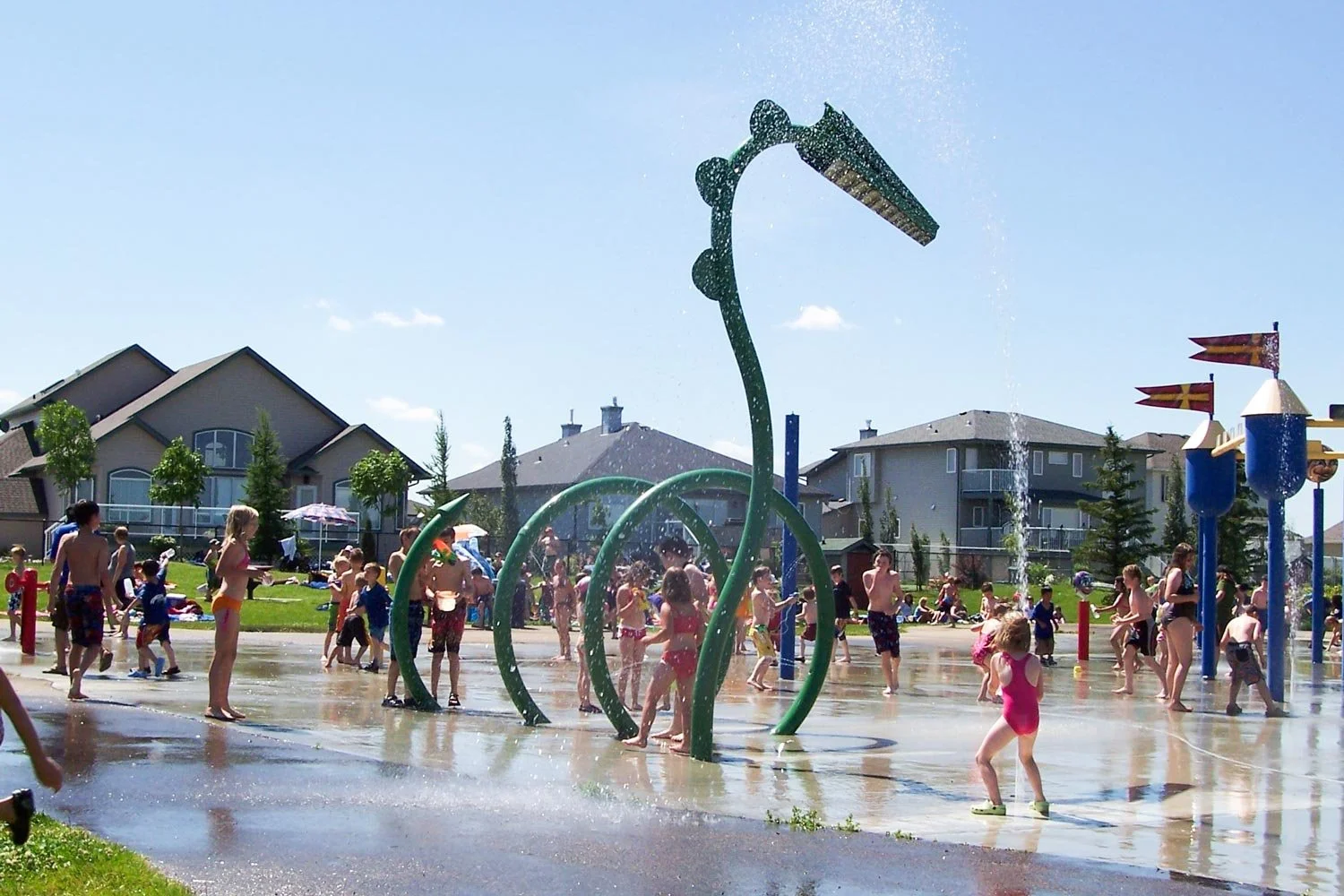 Kids and adults playing under water sprays in a splash pad with a dinosaur-shaped water fountain, residential houses in the background, and a clear blue sky.
