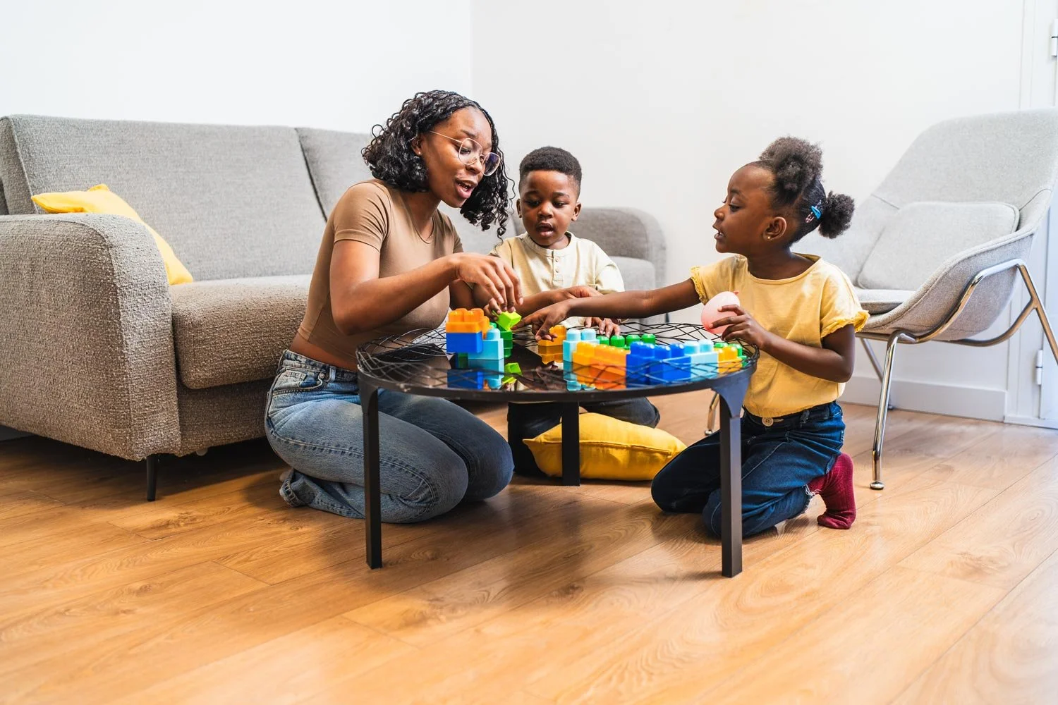 A woman and two young children playing a colorful block stacking game on a small black table in a living room with wooden flooring and gray sofa.