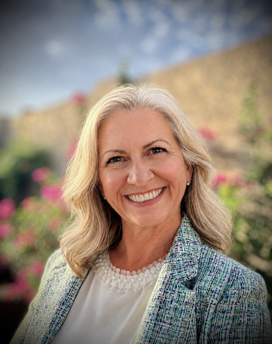 Portrait of a smiling middle-aged woman with blonde wavy hair, wearing a patterned blazer and white blouse, standing outdoors with blurred flowers and greenery in the background.