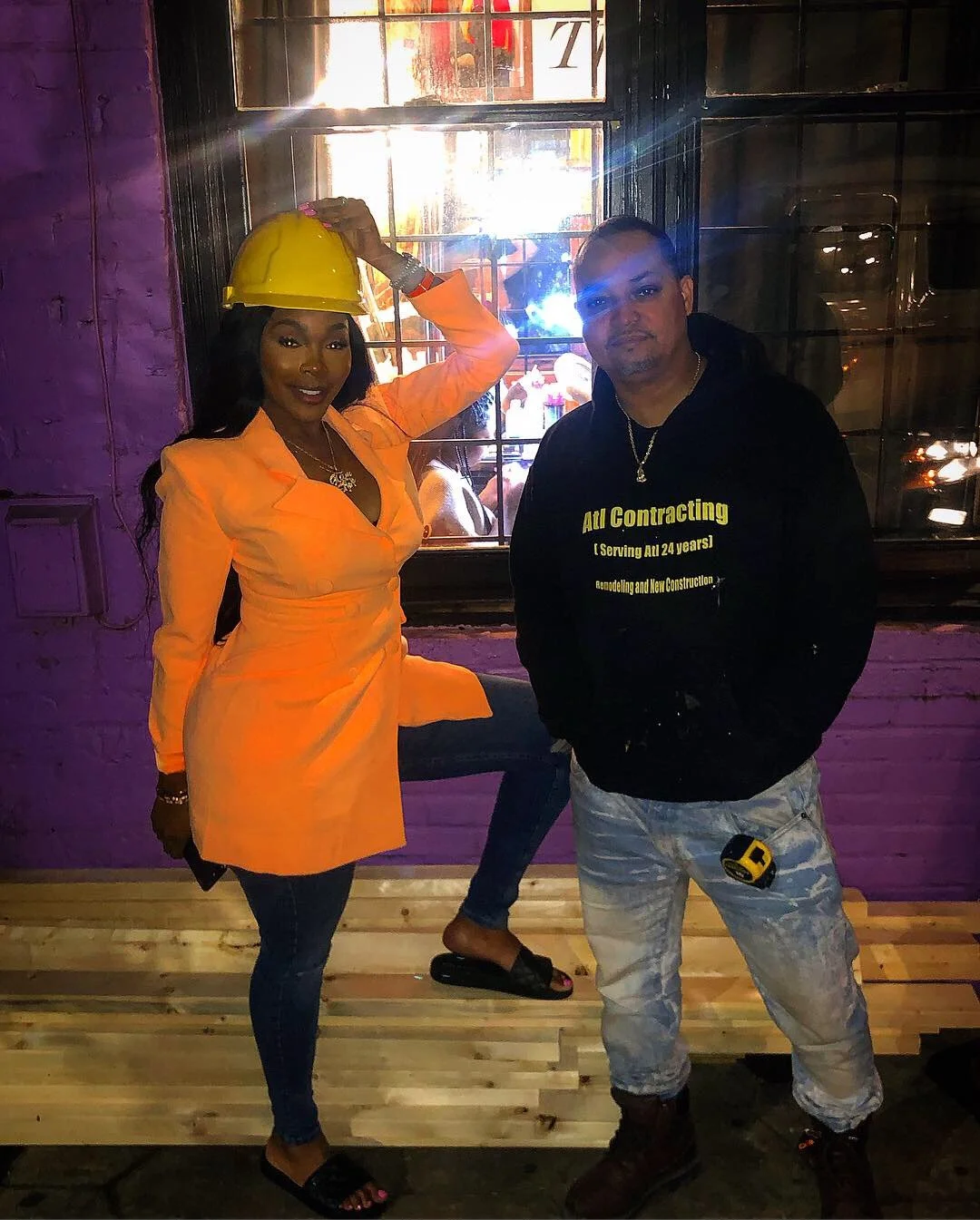 Woman wearing orange construction uniform and yellow hard hat poses with man in black hoodie and camouflage pants inside a building with purple walls and a window.