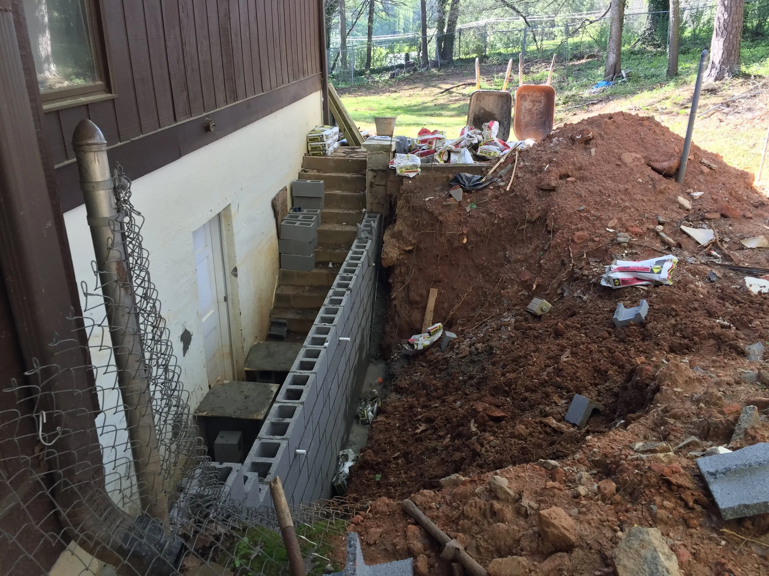 Construction site showing dirt excavation, cinder blocks, a wheelbarrow, and scaffolding against the exterior wall of a house with a forested backyard.