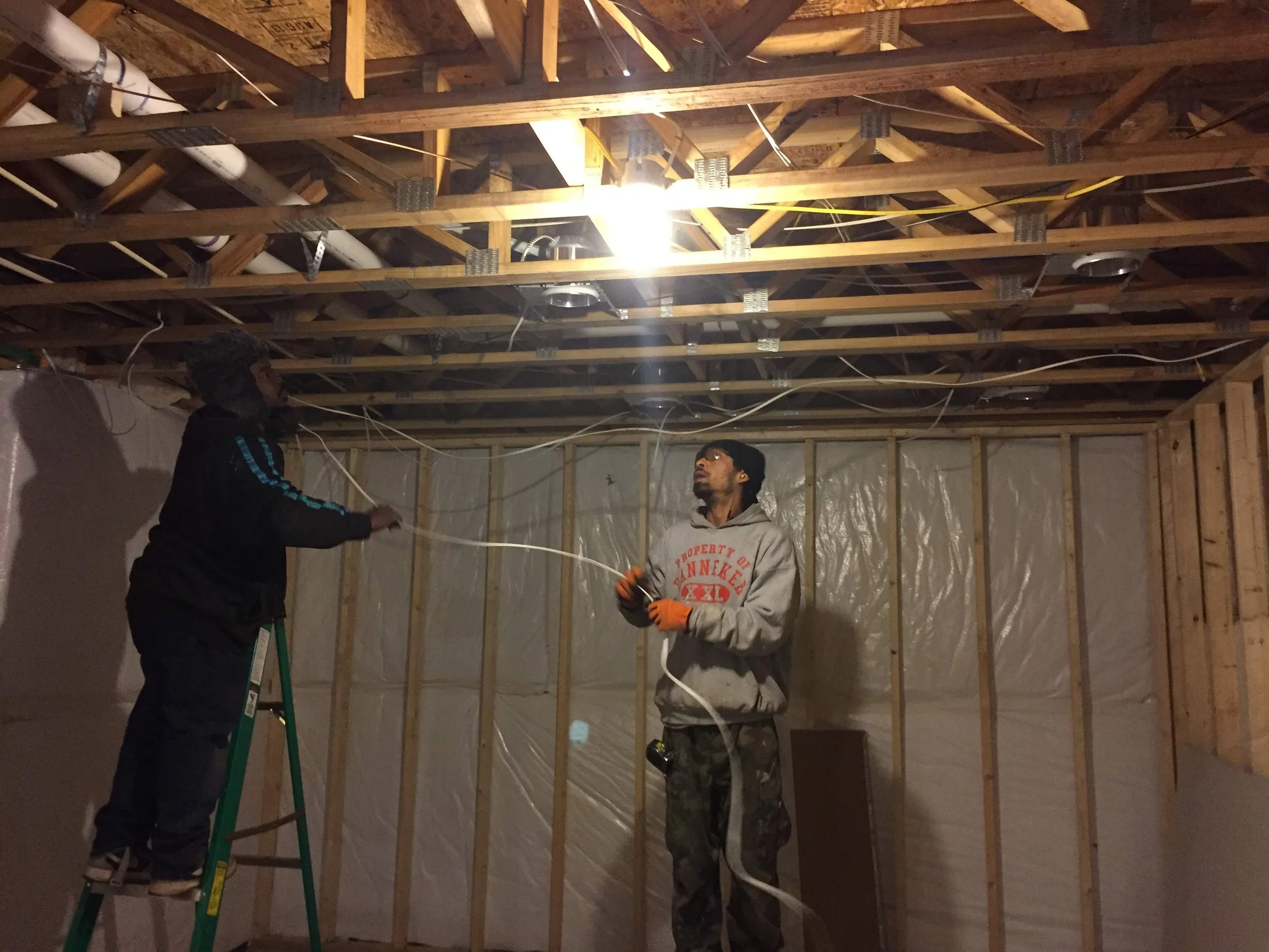 Two men working on electrical wiring and installing ceiling fixtures in an unfinished room with exposed wooden beams and framing.