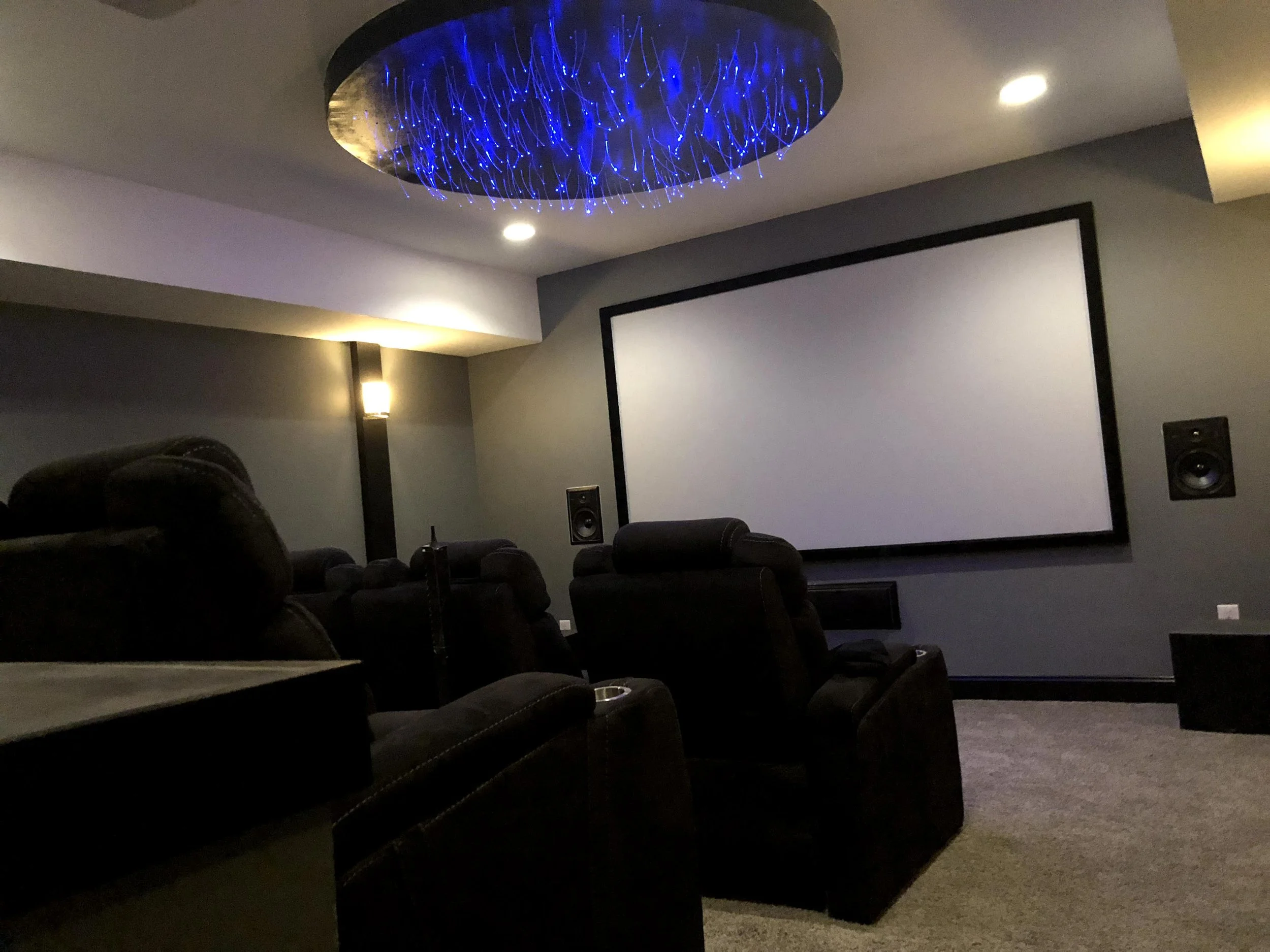 A home theater room with a large projection screen on the wall, black leather recliner chairs, ambient lighting, and a ceiling light fixture with blue LED lights.