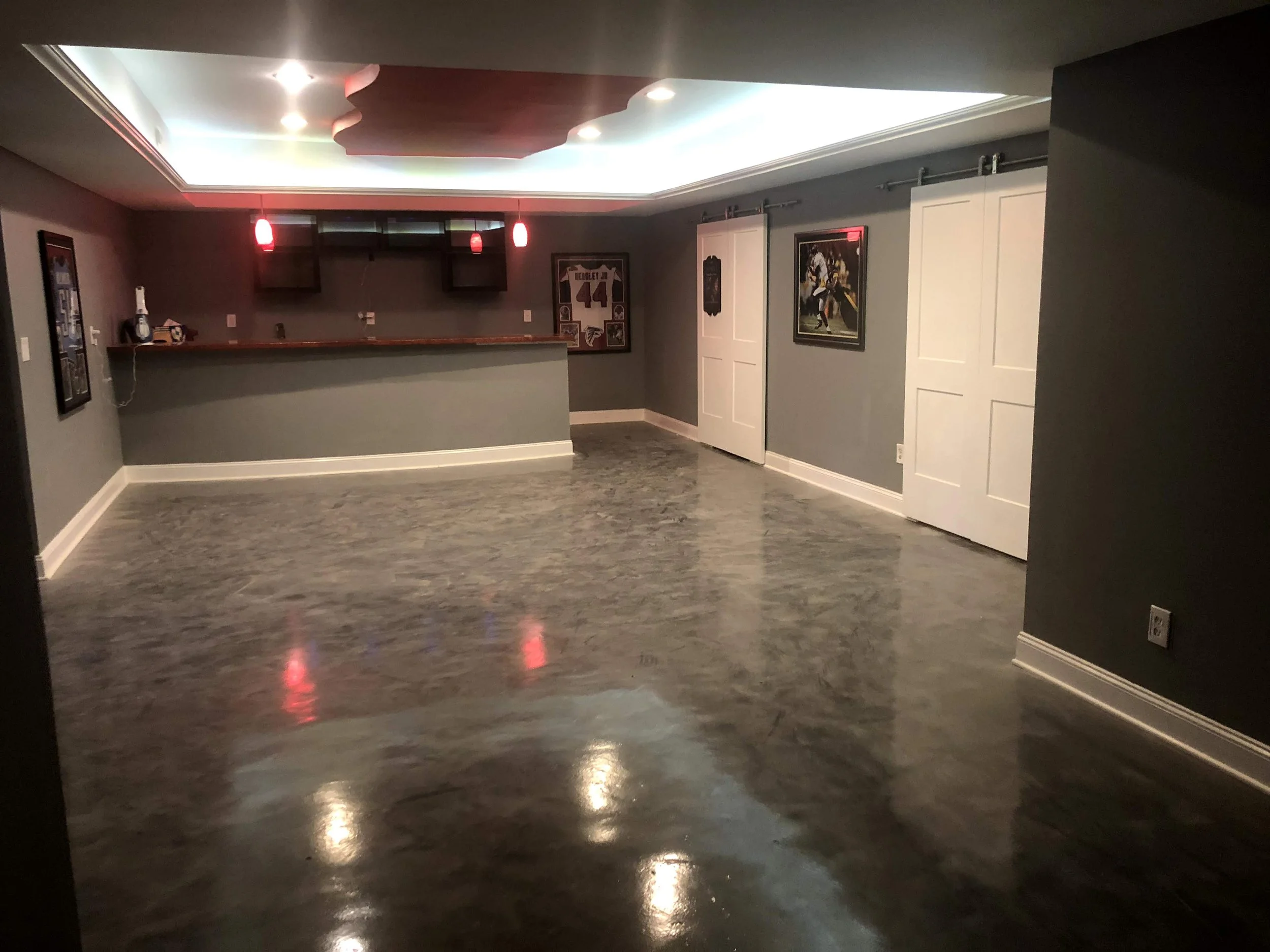 Empty basement room with gray walls, wooden floor, behind a bar counter with hanging red lights, and framed sports memorabilia and artwork on the walls.