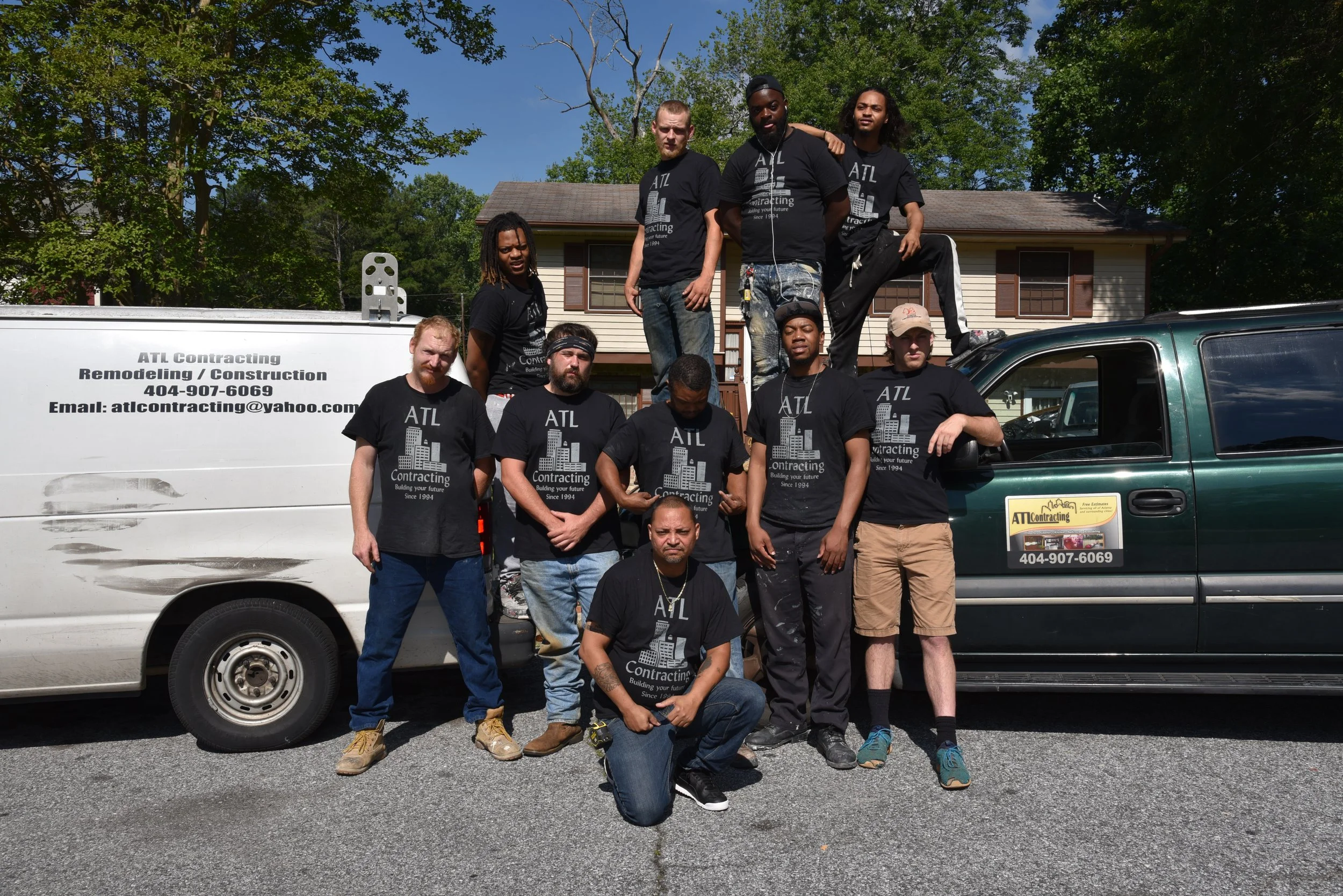 A group of ten men posing in front of a utility van and a green car, with a house and trees in the background, on a sunny day.