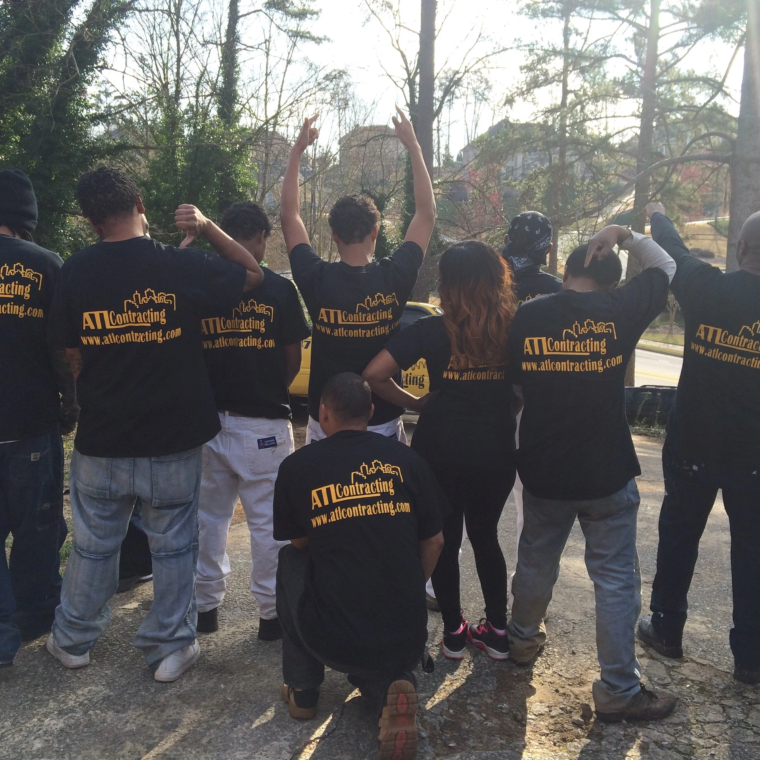 Contracting company, with some raising their hands and others holding a person in a handstand, wearing matching black ATL Basement Solutions T-shirts with yellow text and website URL, during daytime.