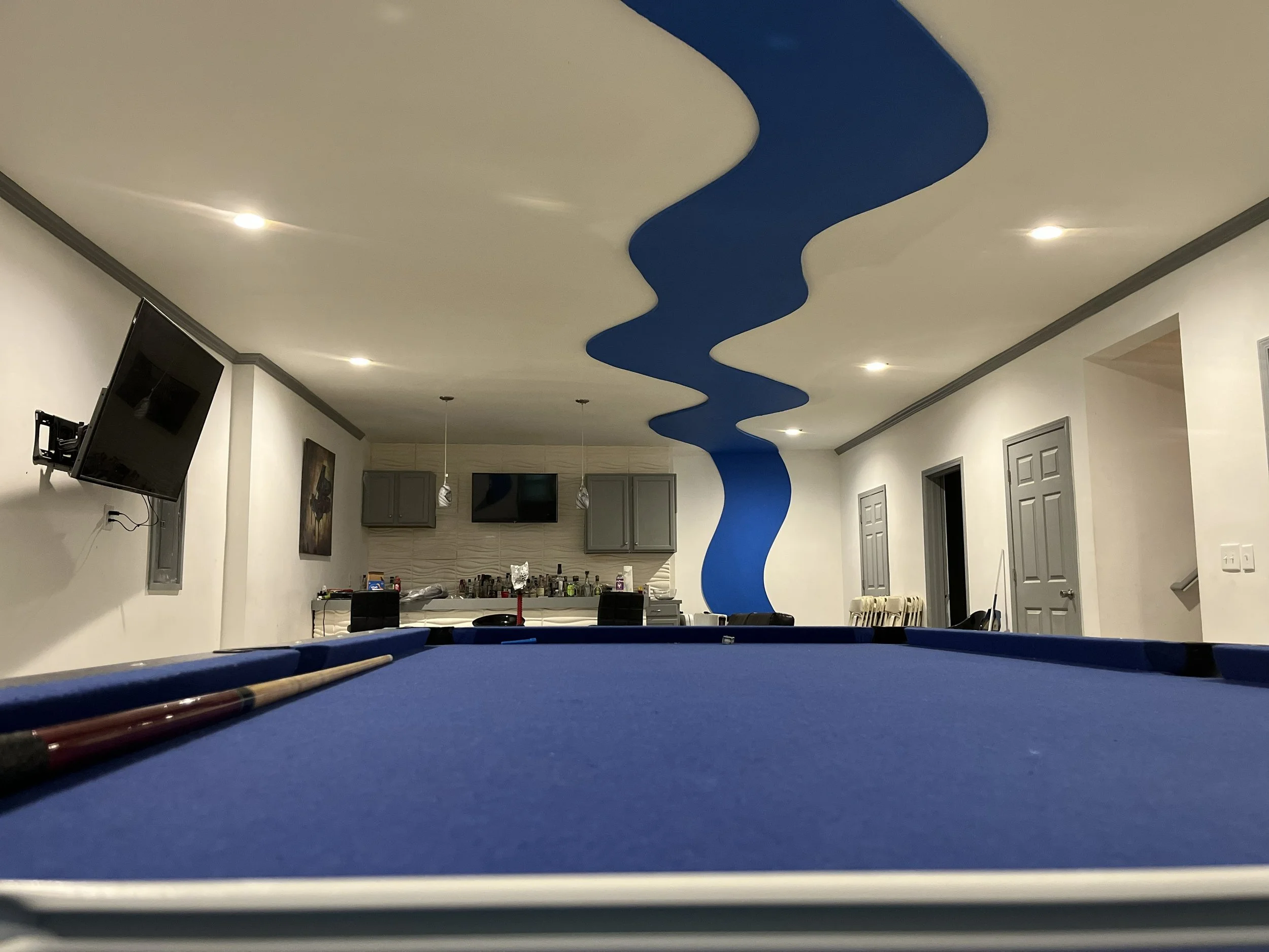 Interior of a room with a blue pool table, pool cues in the foreground, a kitchen area in the background, a blue wavy ceiling design, pendant lights, and wall-mounted televisions. Palmetto Ga.
