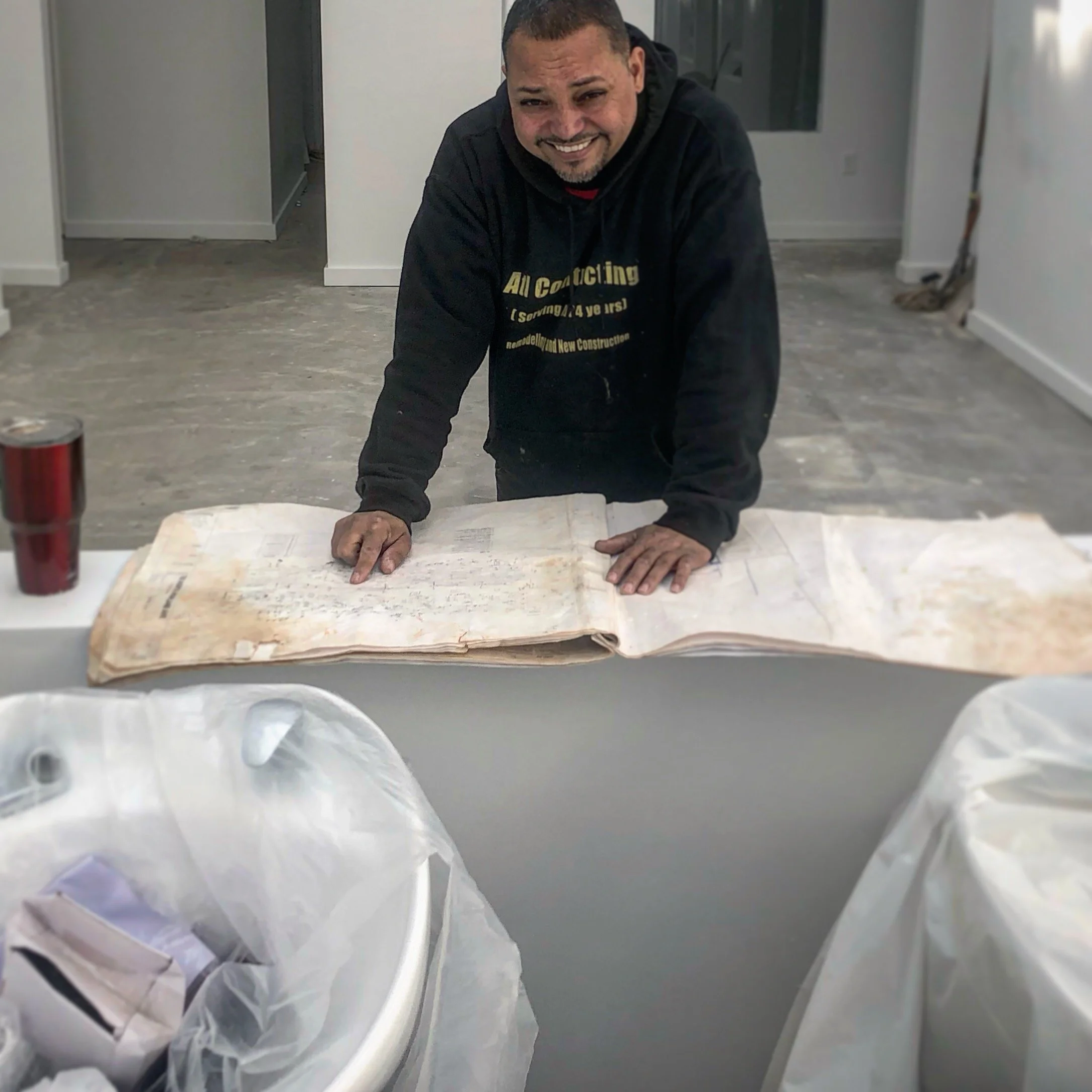 Man smiling while kneeling on the floor in a room under renovation, with crumpled paper and construction materials around him.