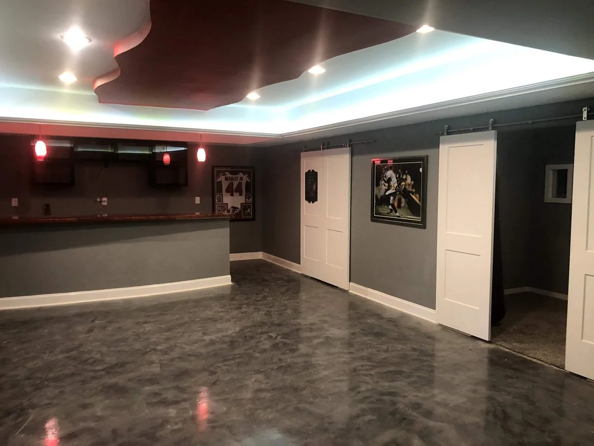 basement room with dark gray walls, white sliding barn doors, a bar area with hanging lights, sports posters on the walls, and polished gray concrete floors.