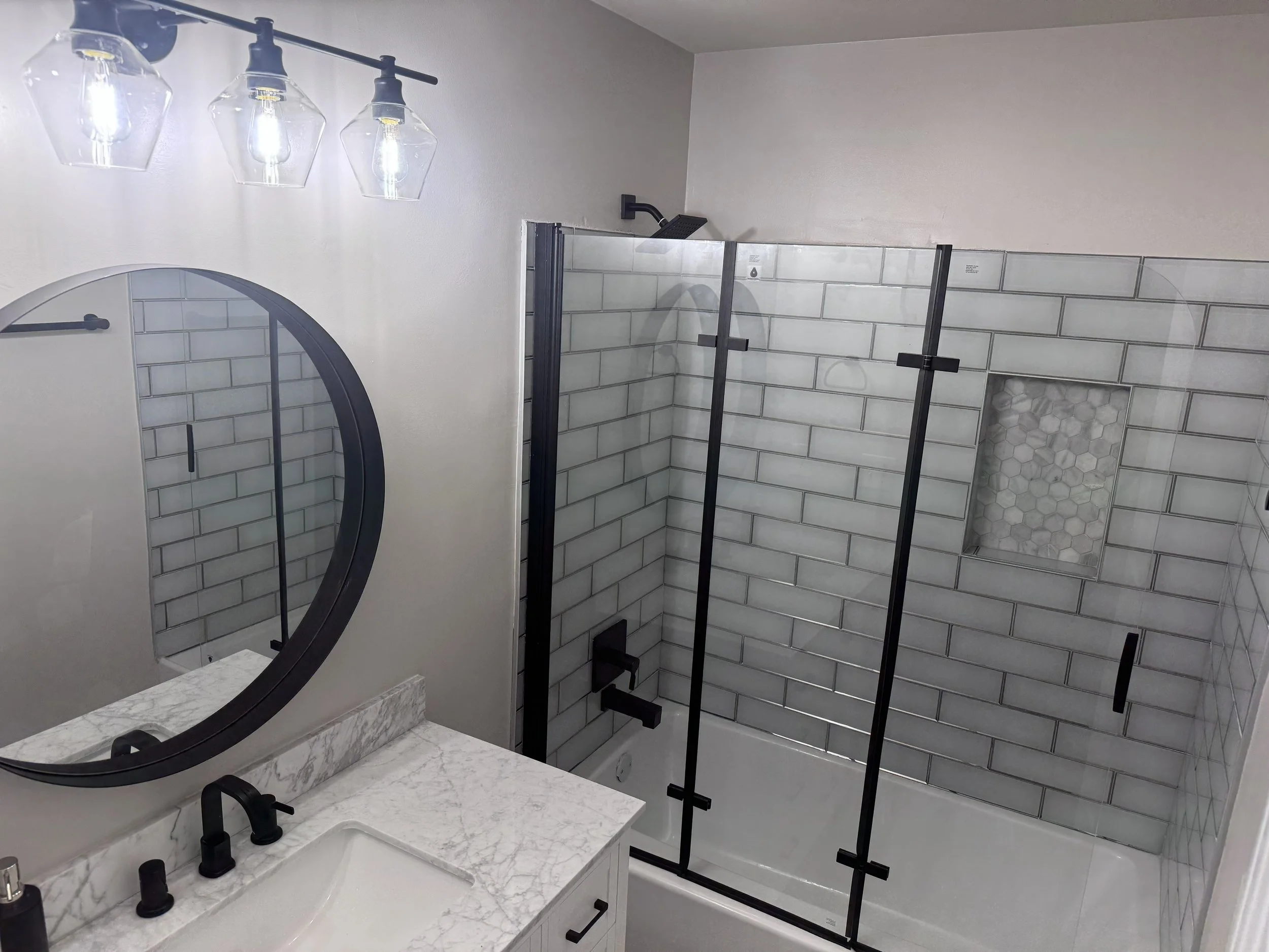 Modern bathroom with a shower enclosed by glass panels, white subway tiles, a circular mirror, and a marble countertop with black fixtures. Decatur Ga.