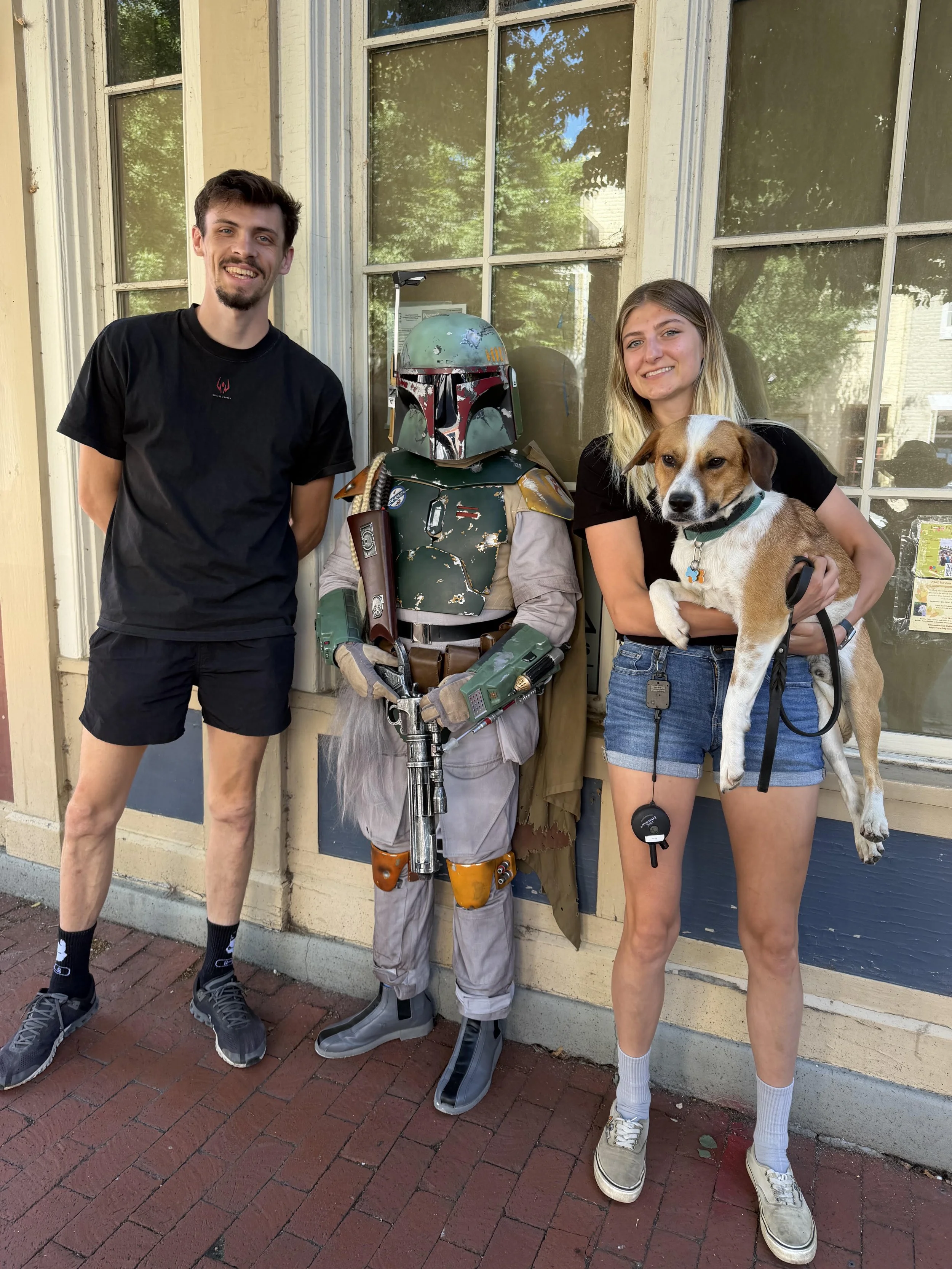 Three young people and a dog standing outside a building with large windows in a historic area.