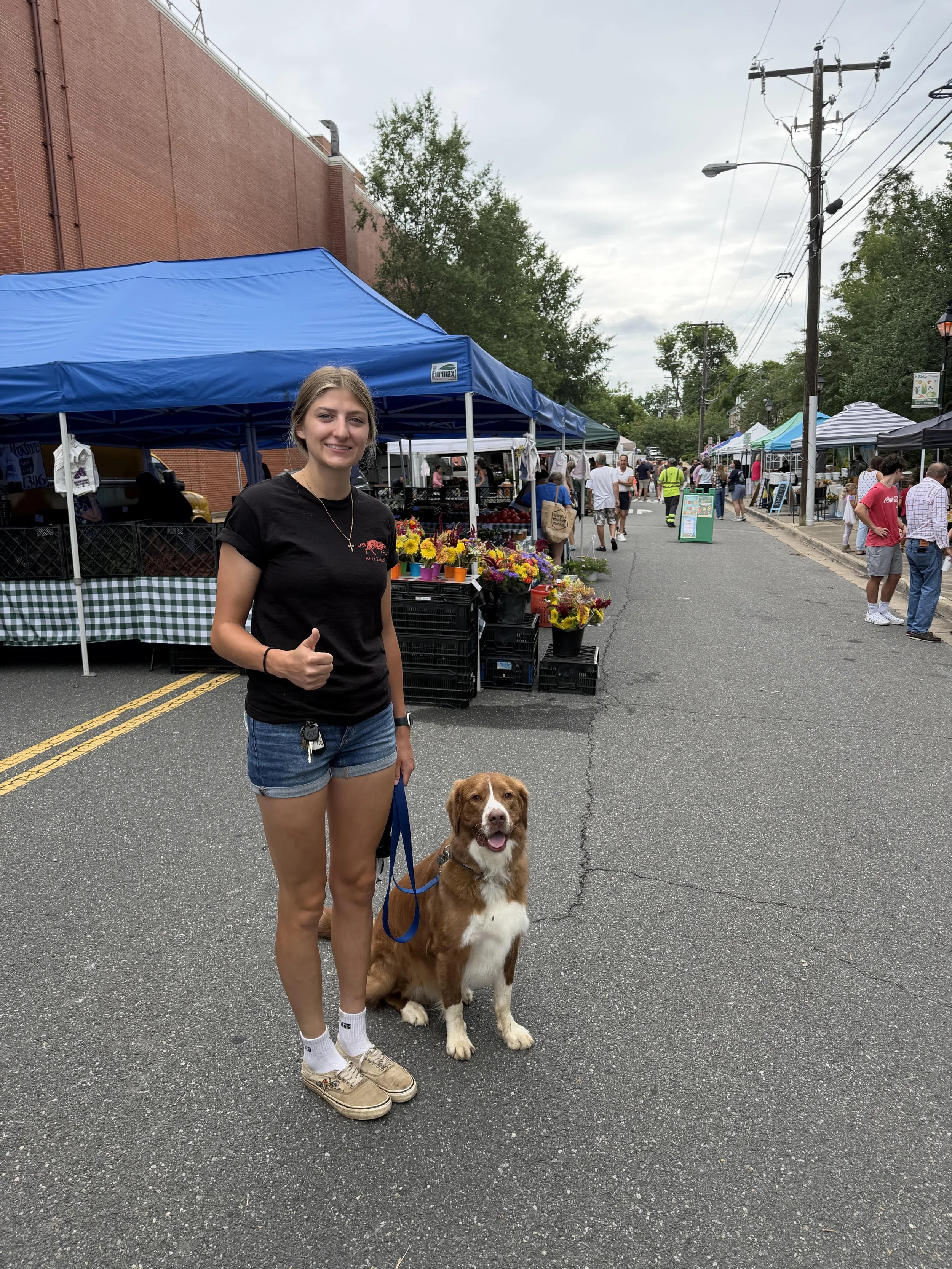 A young woman standing on a street fair with a brown and white dog on a leash. She is giving a thumbs-up and smiling. There are tents and people browsing in the background on a cloudy day.